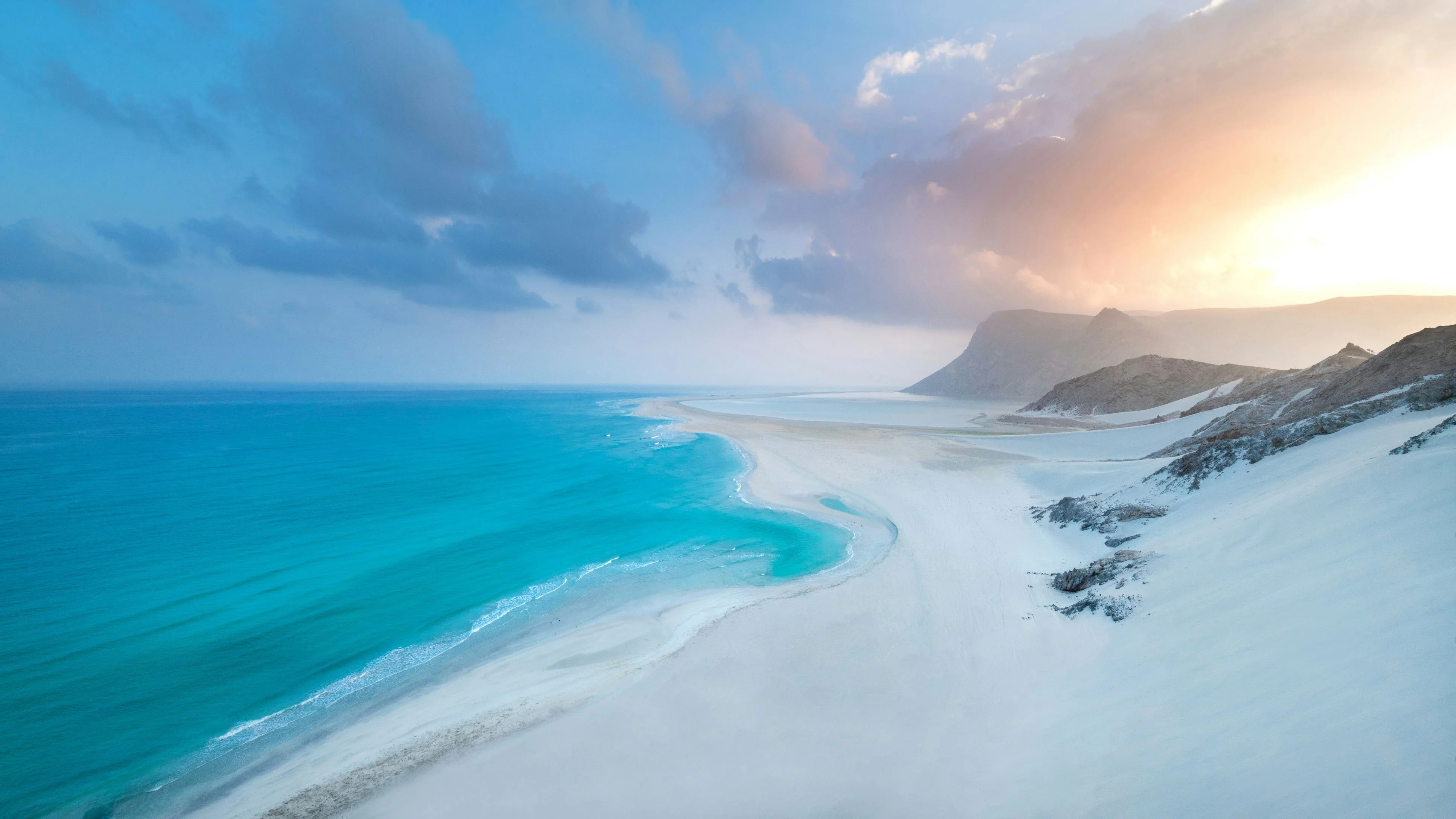 Aerial view of white sand dunes meeting turquoise lagoon and mountains on Socotra at dawn.
