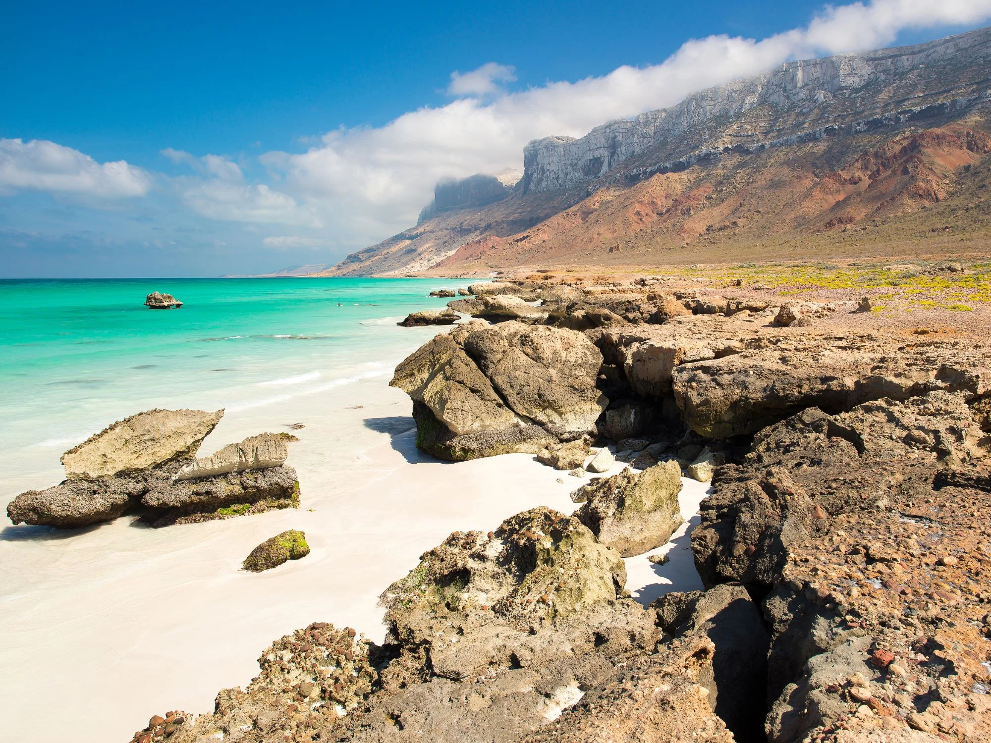 Turquoise Socotra beach with white sand, rock formations and towering cliffs under blue skies.