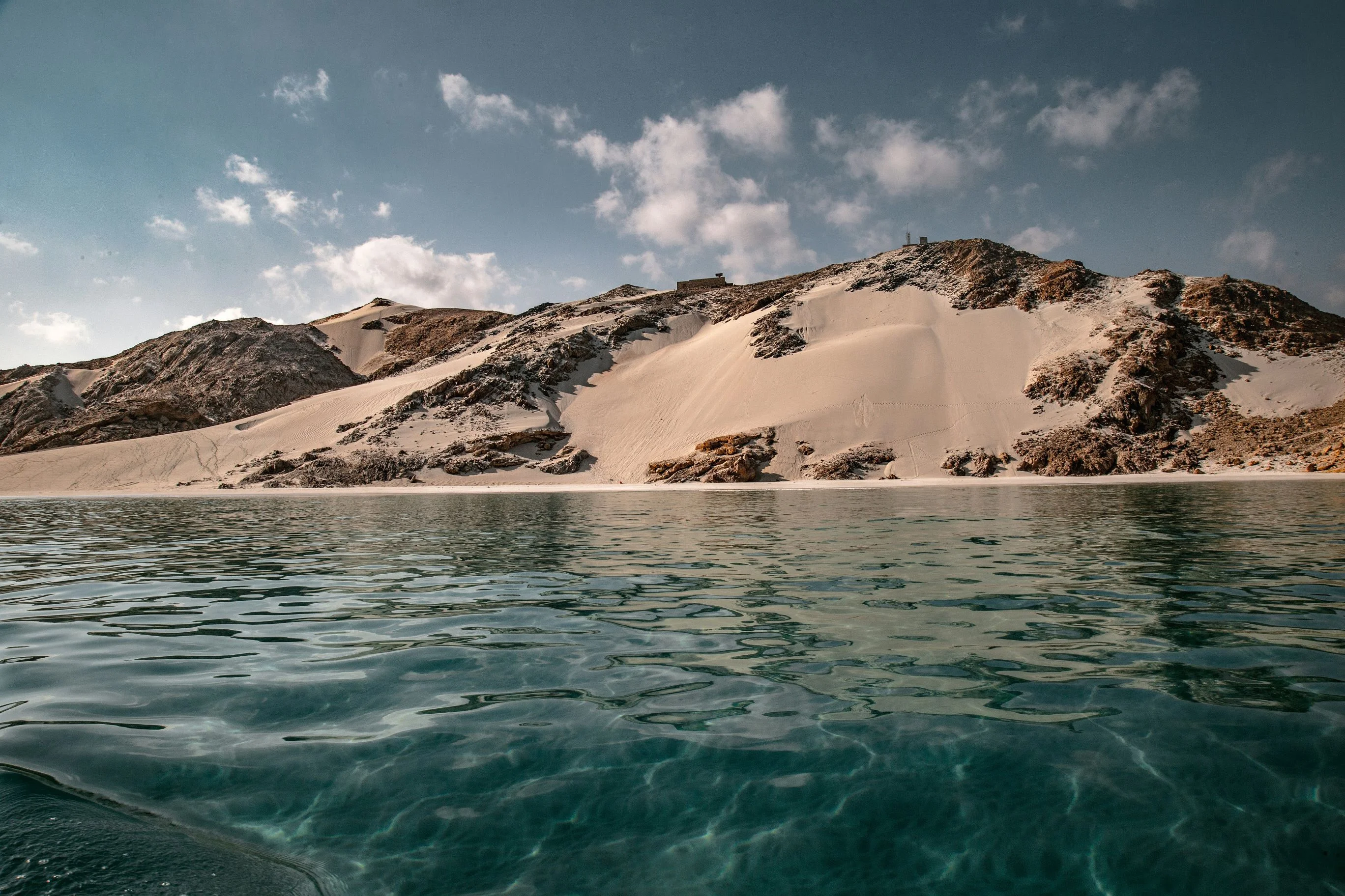 Turquoise waters lap against sand dune and rocky cliffs on Socotra under blue skies.