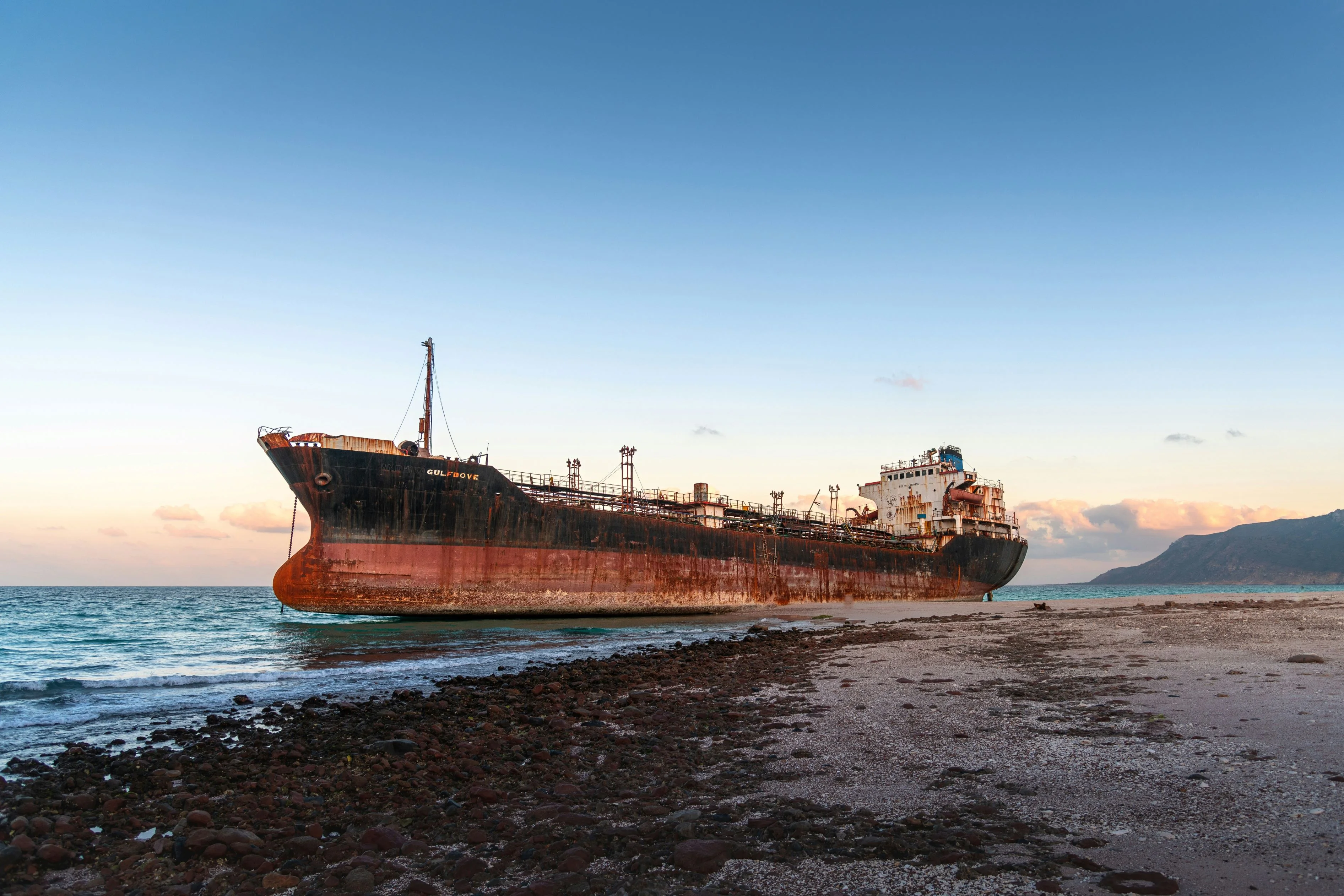 Rusting cargo shipwreck stranded on Socotra shore with turquoise sea and mountains in background.