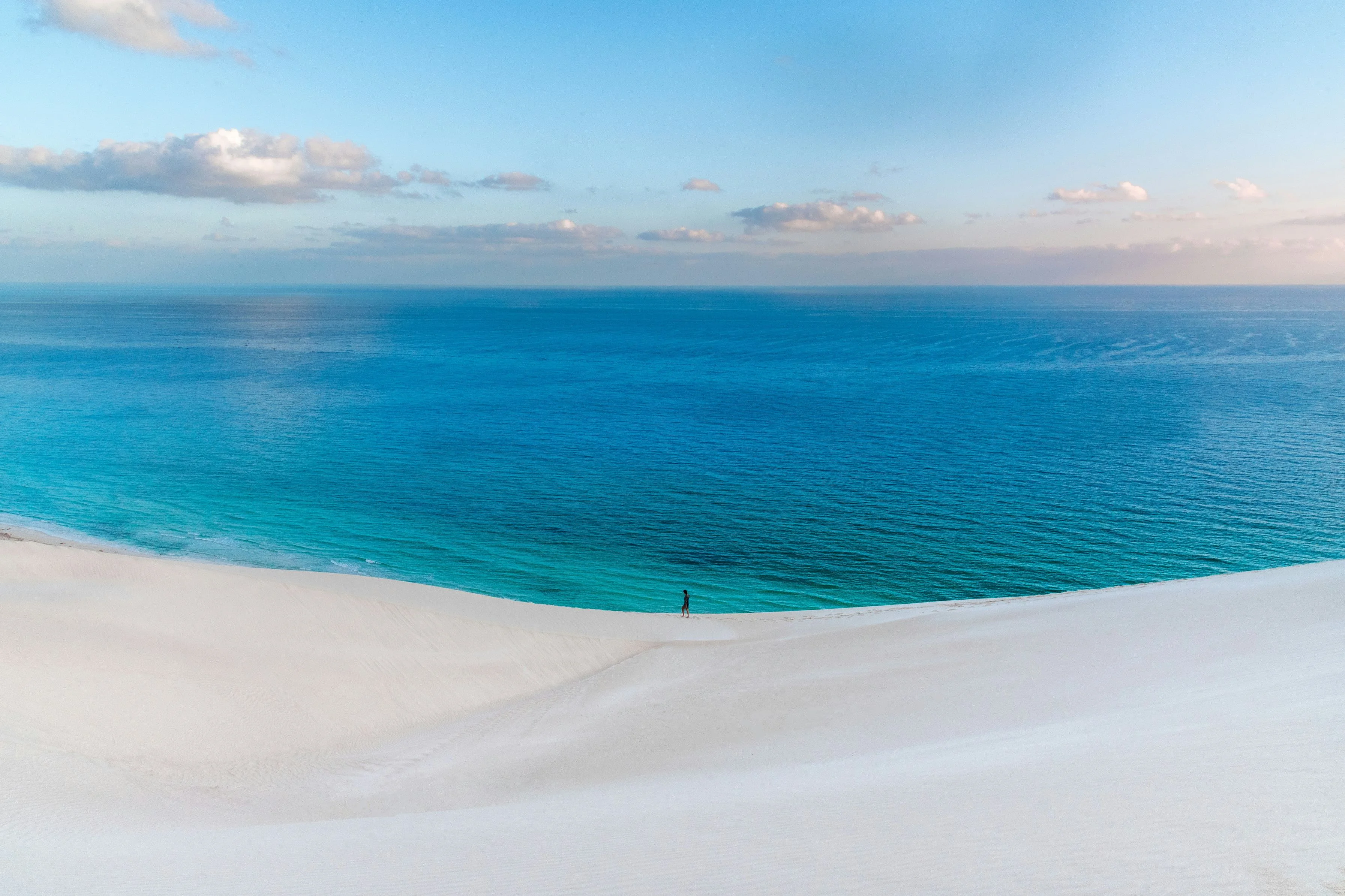 Solitary person atop vast white dune overlooking deep blue Arabian Sea on Socotra.