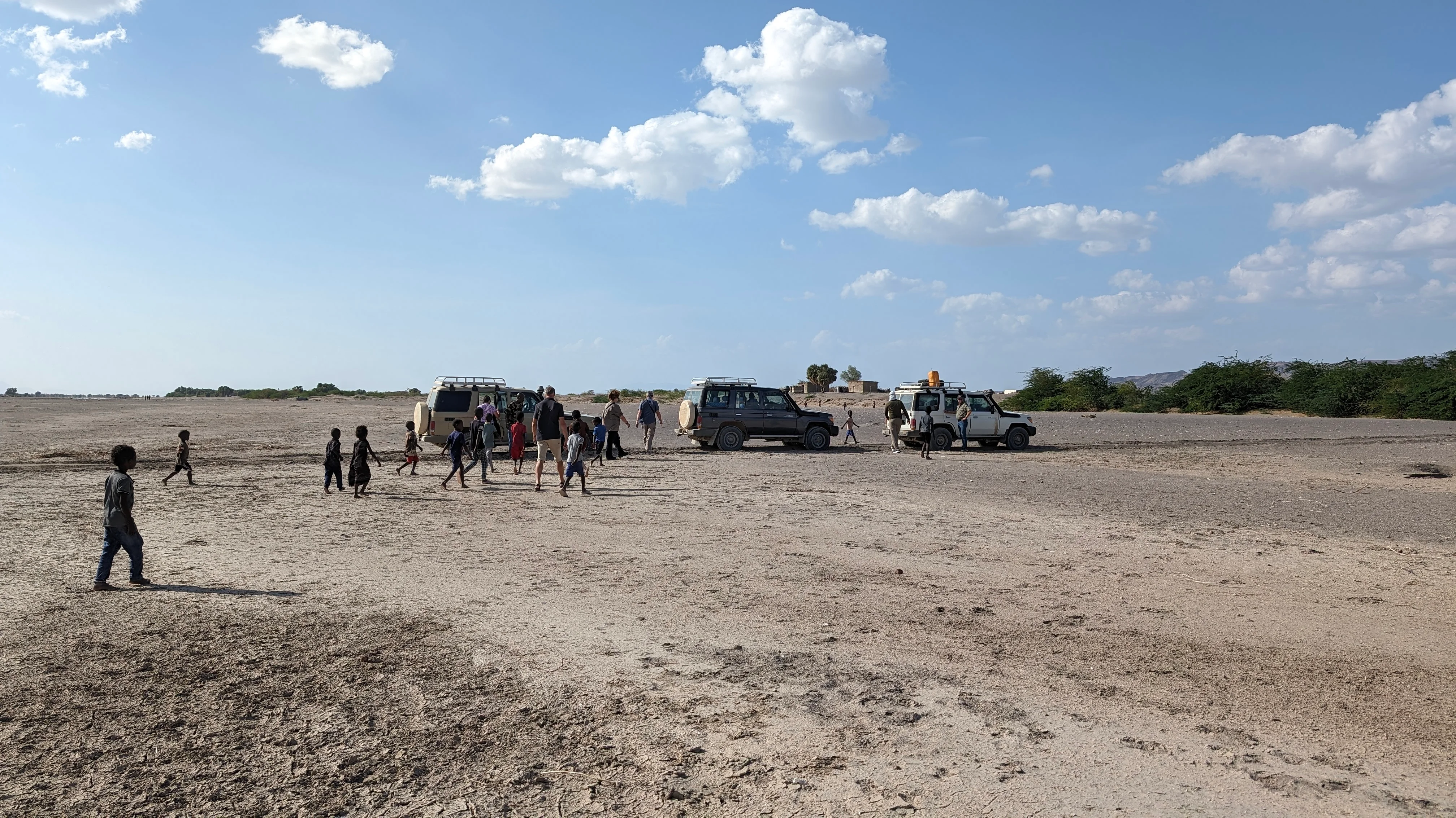 Convoy of 4x4 vehicles parked on barren plain as local children approach under blue sky.