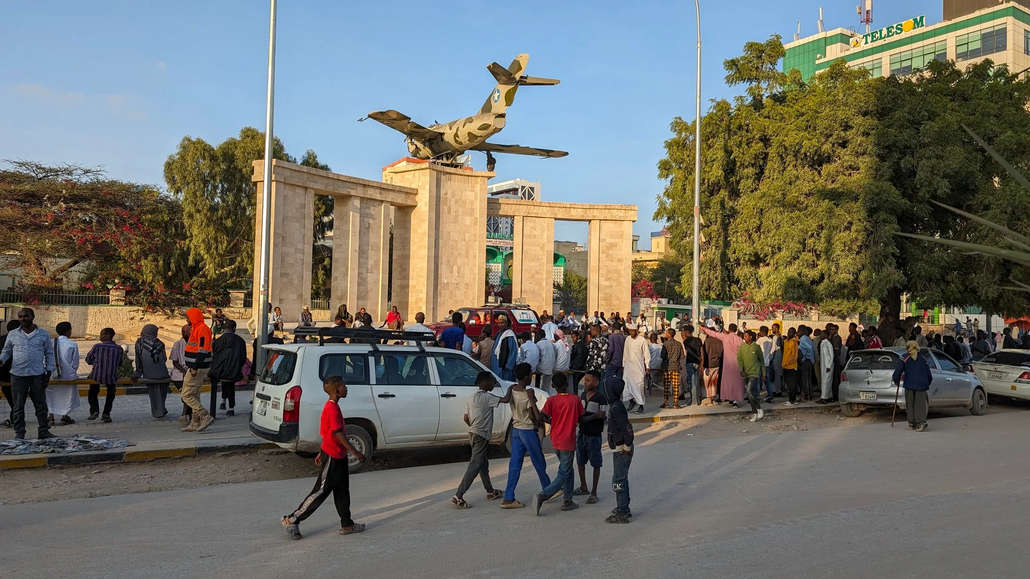 Crowd gathers at Hargeisa war memorial with downed fighter jet monument and flags in Somaliland.