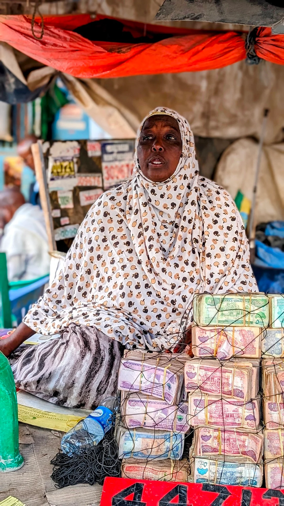 Hijab-wearing woman sits behind bundles of colorful Somaliland shilling notes at street money changer.