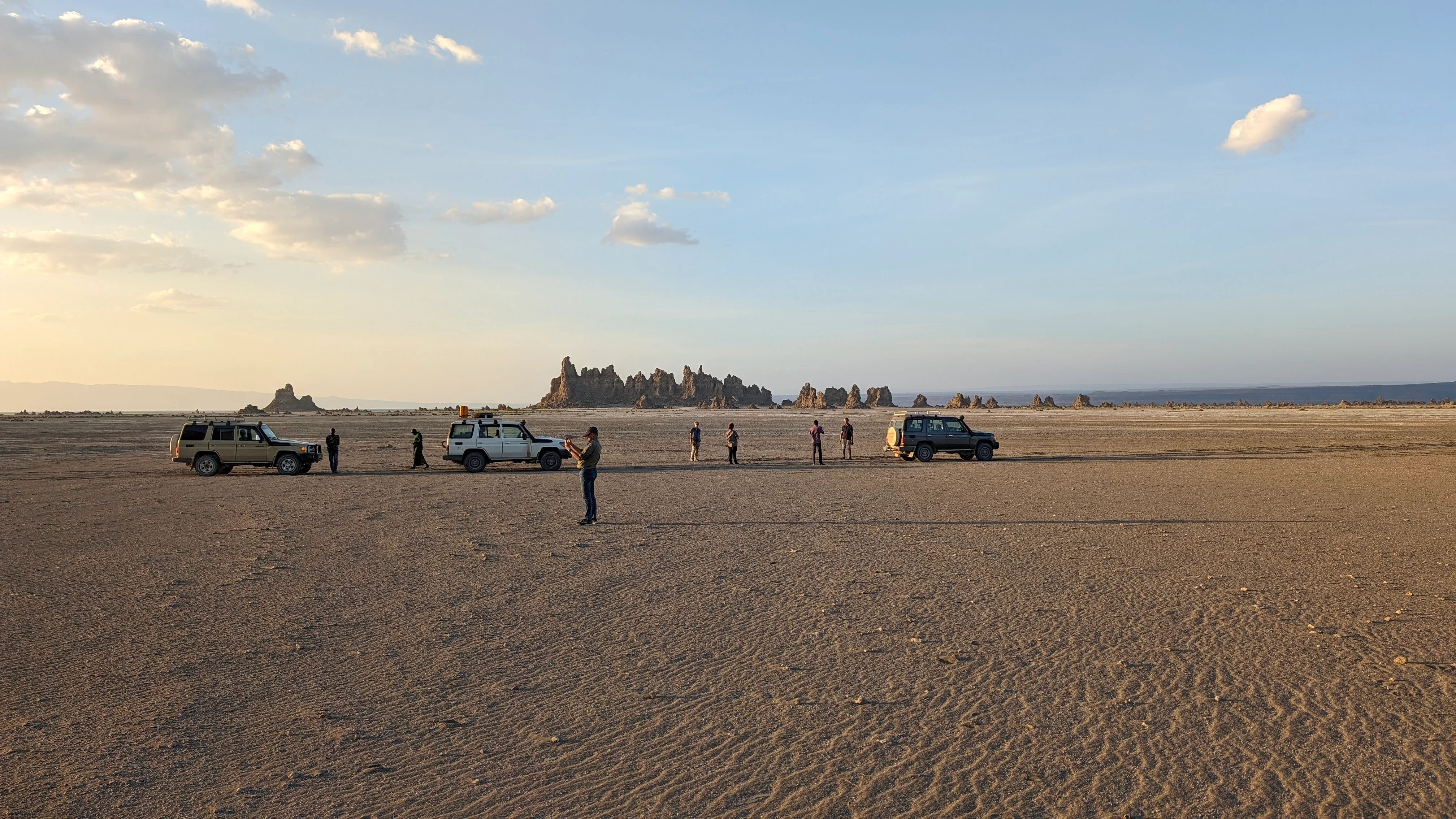 Safari vehicles and travellers on open salt flats of Lake Abbe with distant limestone chimneys.
