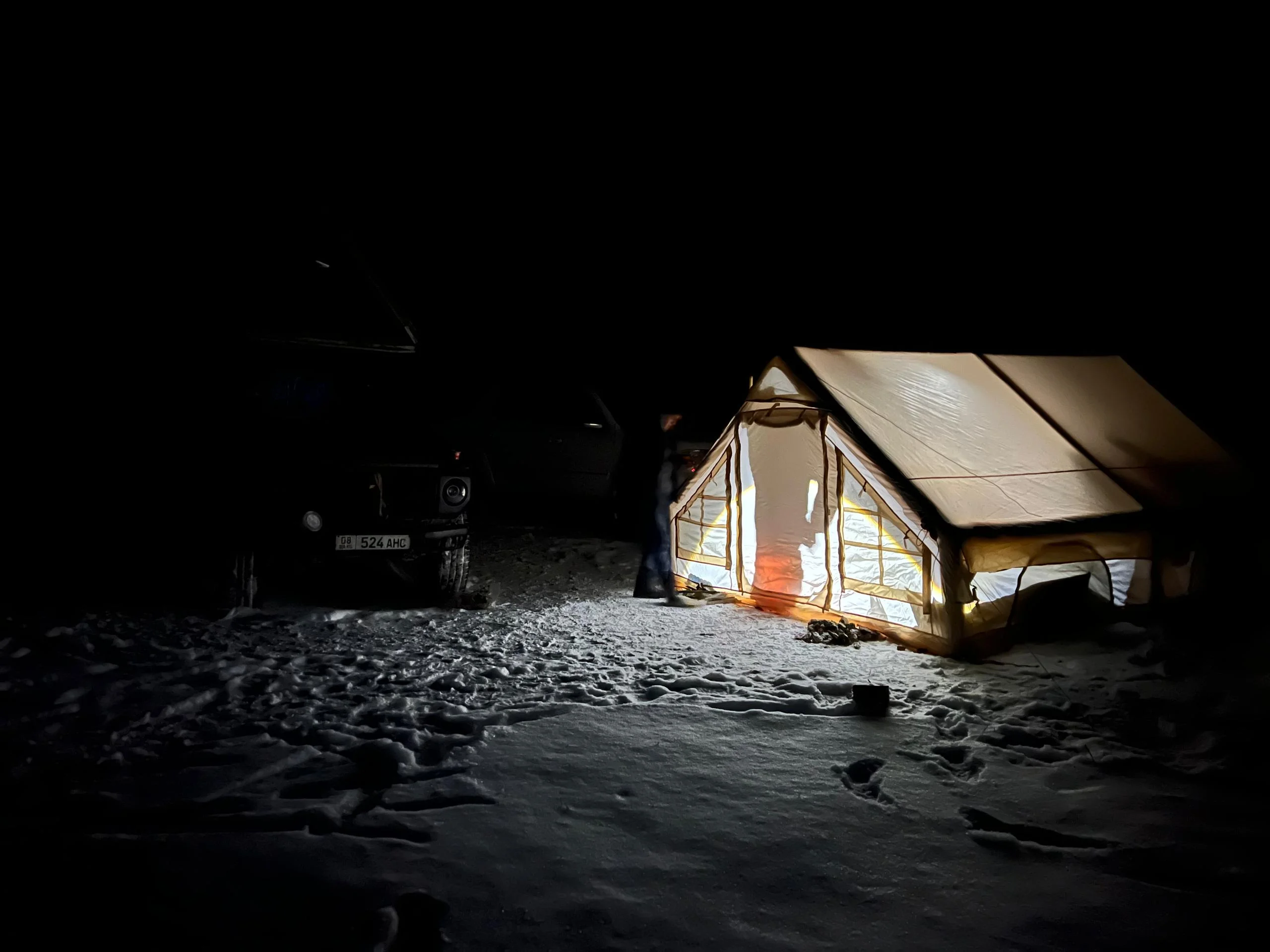 Illuminated canvas expedition tent glowing warmly beside a 4x4 vehicle in a dark snowy winter camp in Kyrgyzstan
