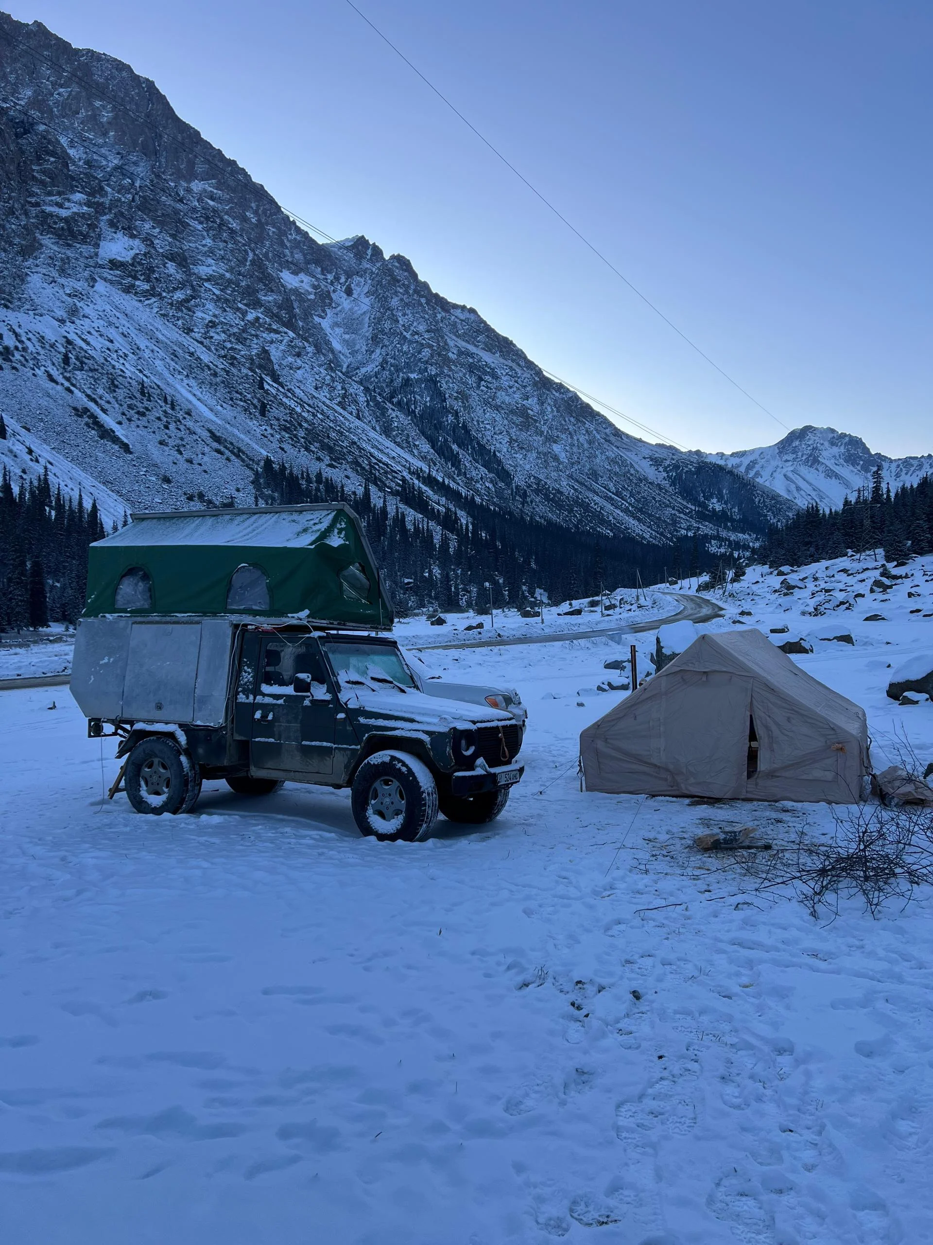 Snow-dusted expedition camp at dawn in a mountain valley with a rugged 4x4 rooftop tent and canvas shelter below steep Tien Shan peaks