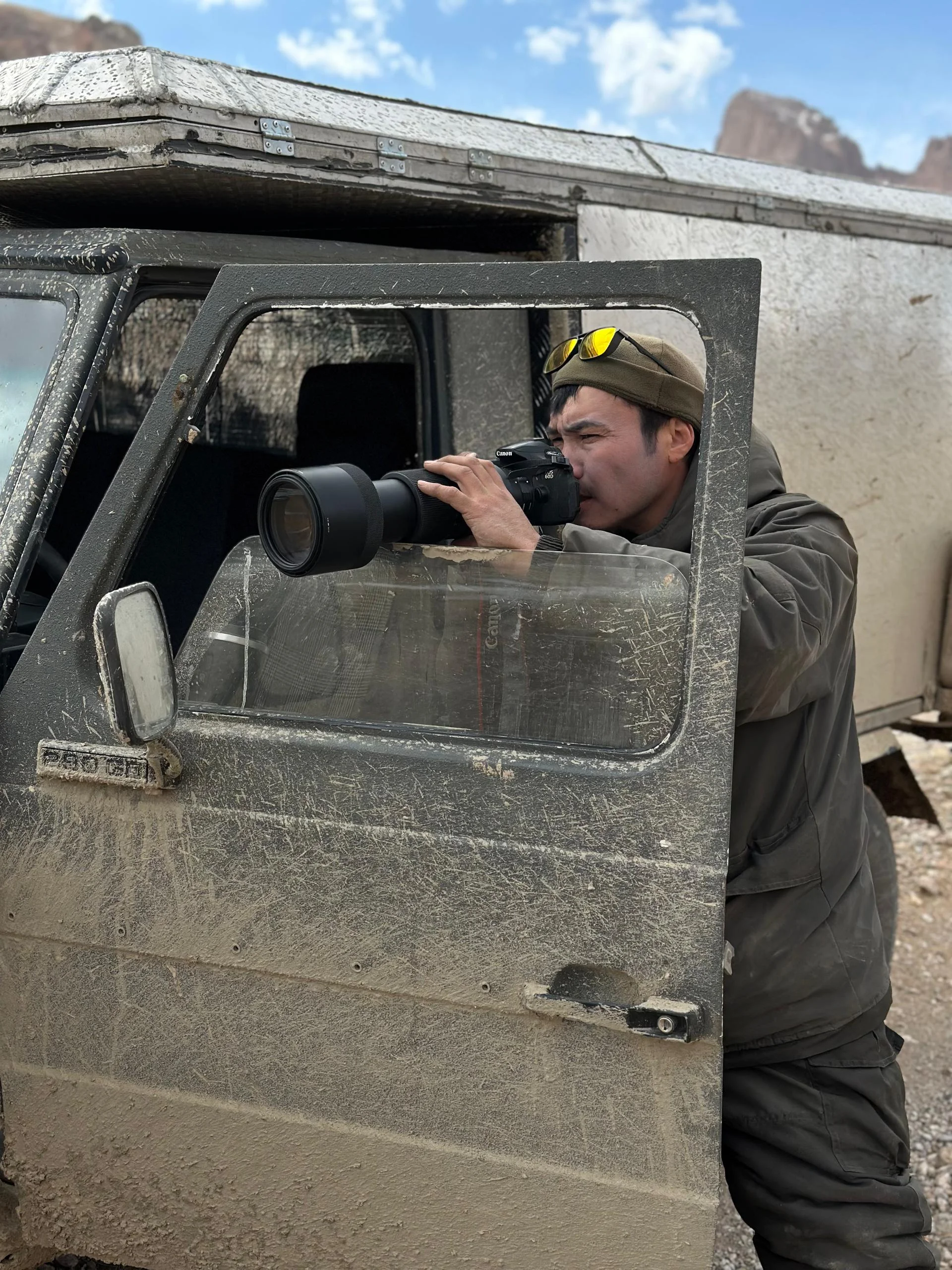 Expedition guide photographing through a Canon telephoto lens from the open door of a dusty 4x4 vehicle in the Kyrgyz mountains