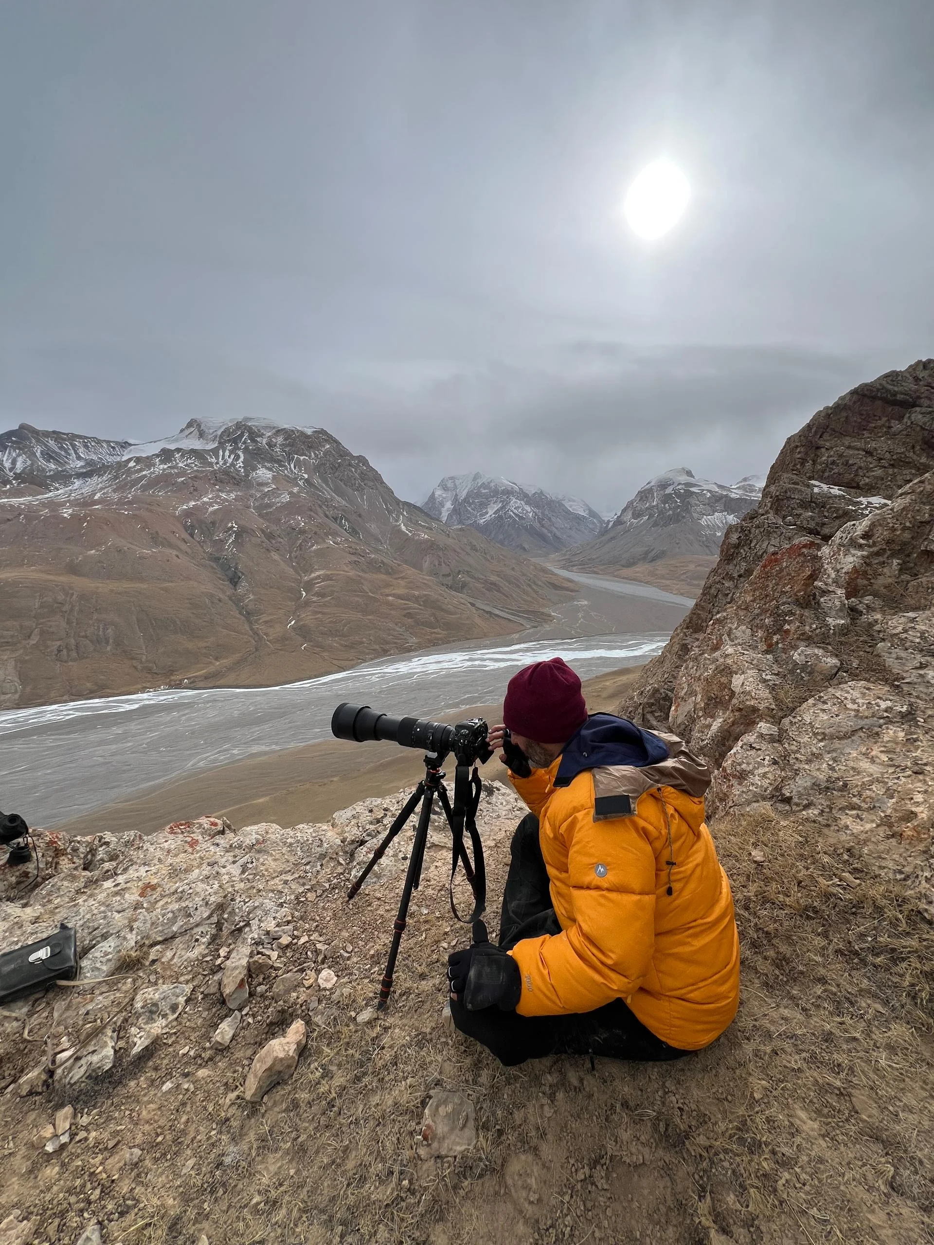 Wildlife photographer in orange down jacket shooting with a telephoto lens on a tripod from a rocky outcrop above a snowy mountain valley
