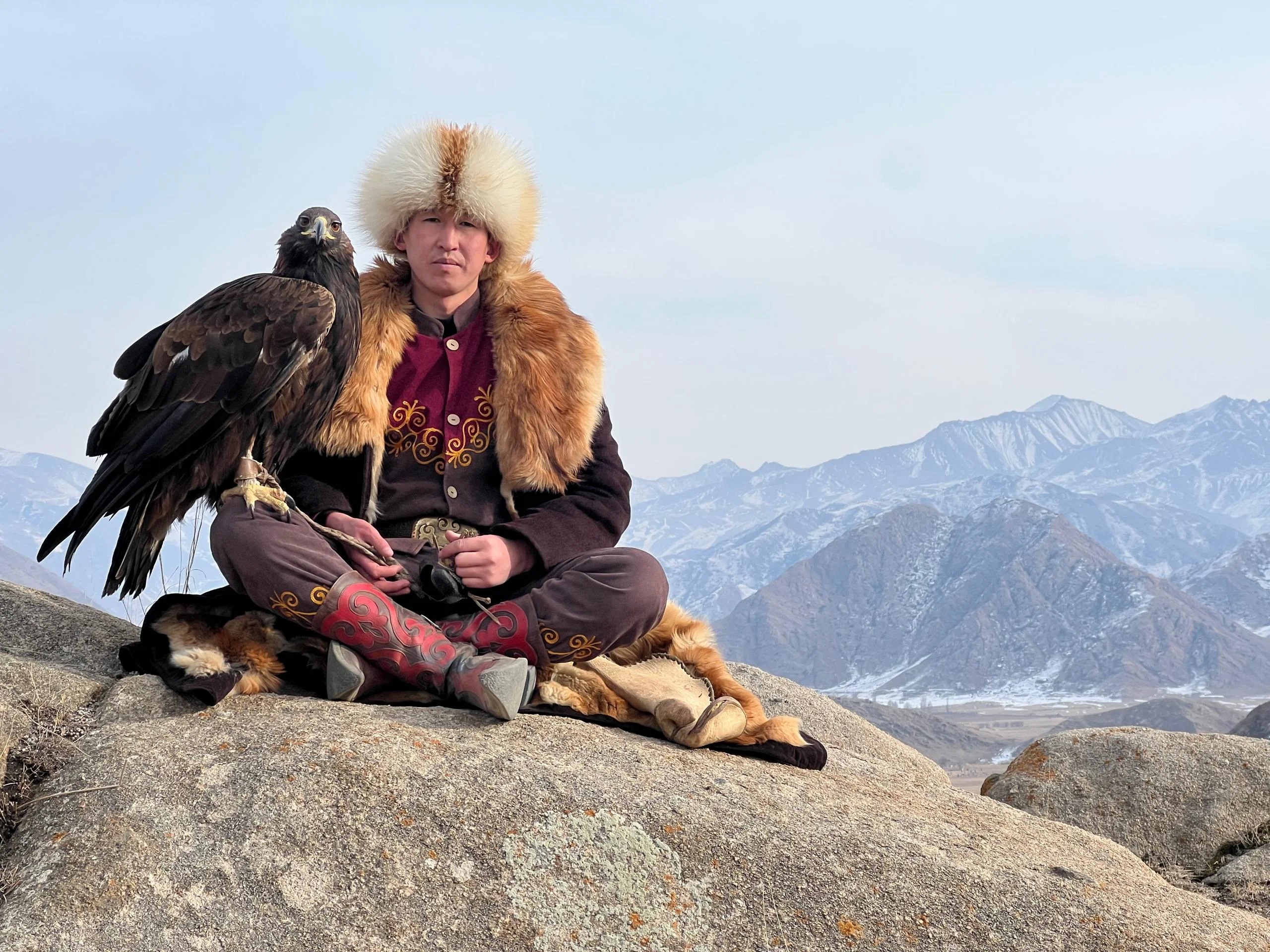Young Kyrgyz eagle hunter in traditional embroidered coat and fur kalpak hat seated on a rock with a golden eagle, mountains behind