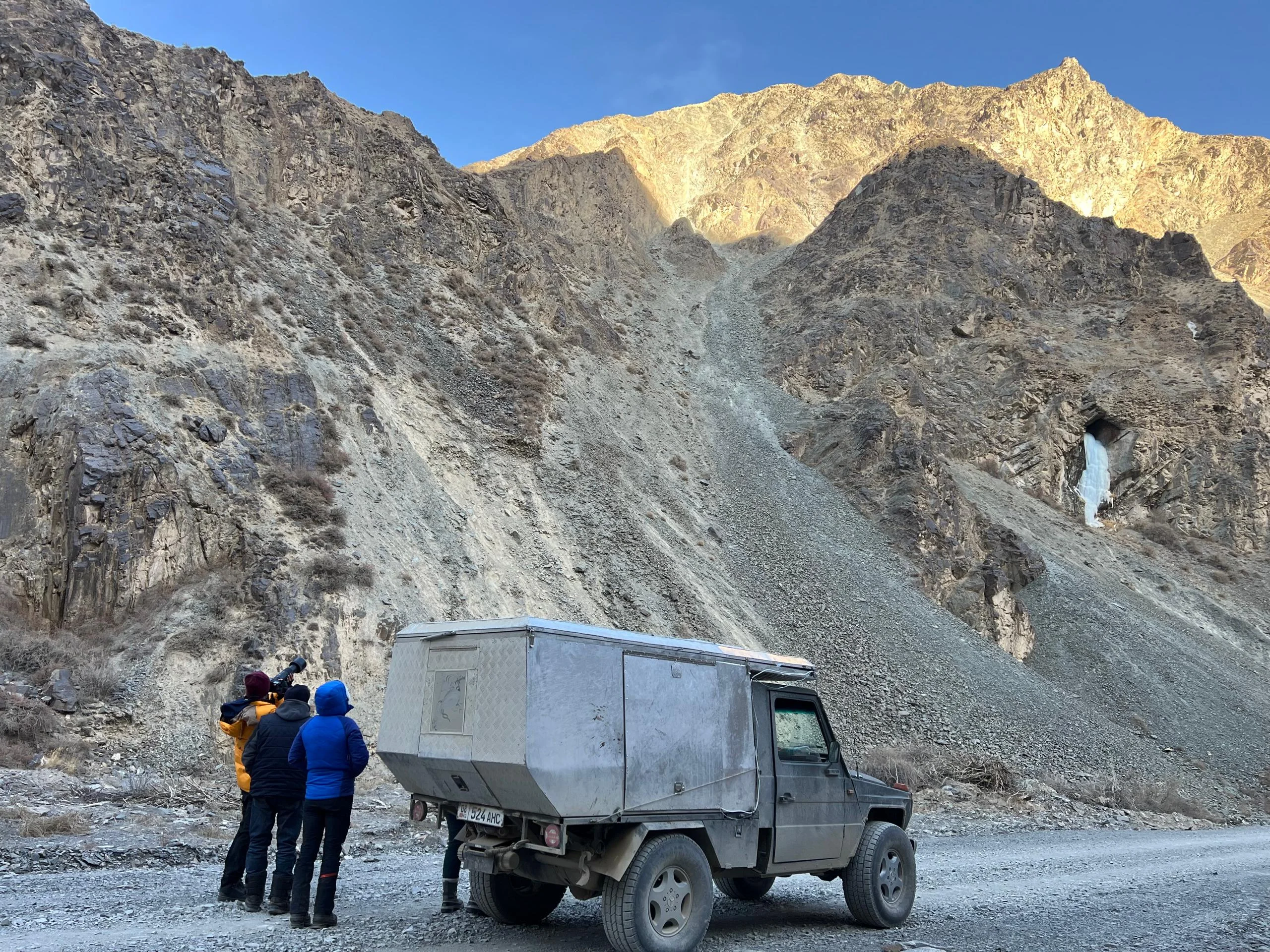 Three expedition members scanning a dramatic rock canyon with a frozen waterfall beside a rugged 4x4 vehicle in the Tien Shan