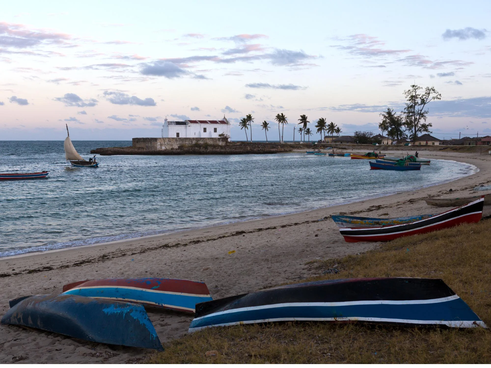 Colorful wooden fishing boats beached near a white colonial chapel on a quiet bay at sunset, Mozambique