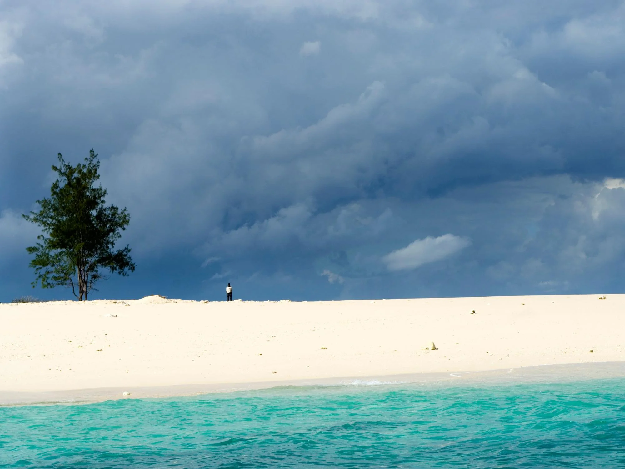 Lone figure standing on a pristine white sand island with a single tree under a dramatic stormy sky, Mozambique