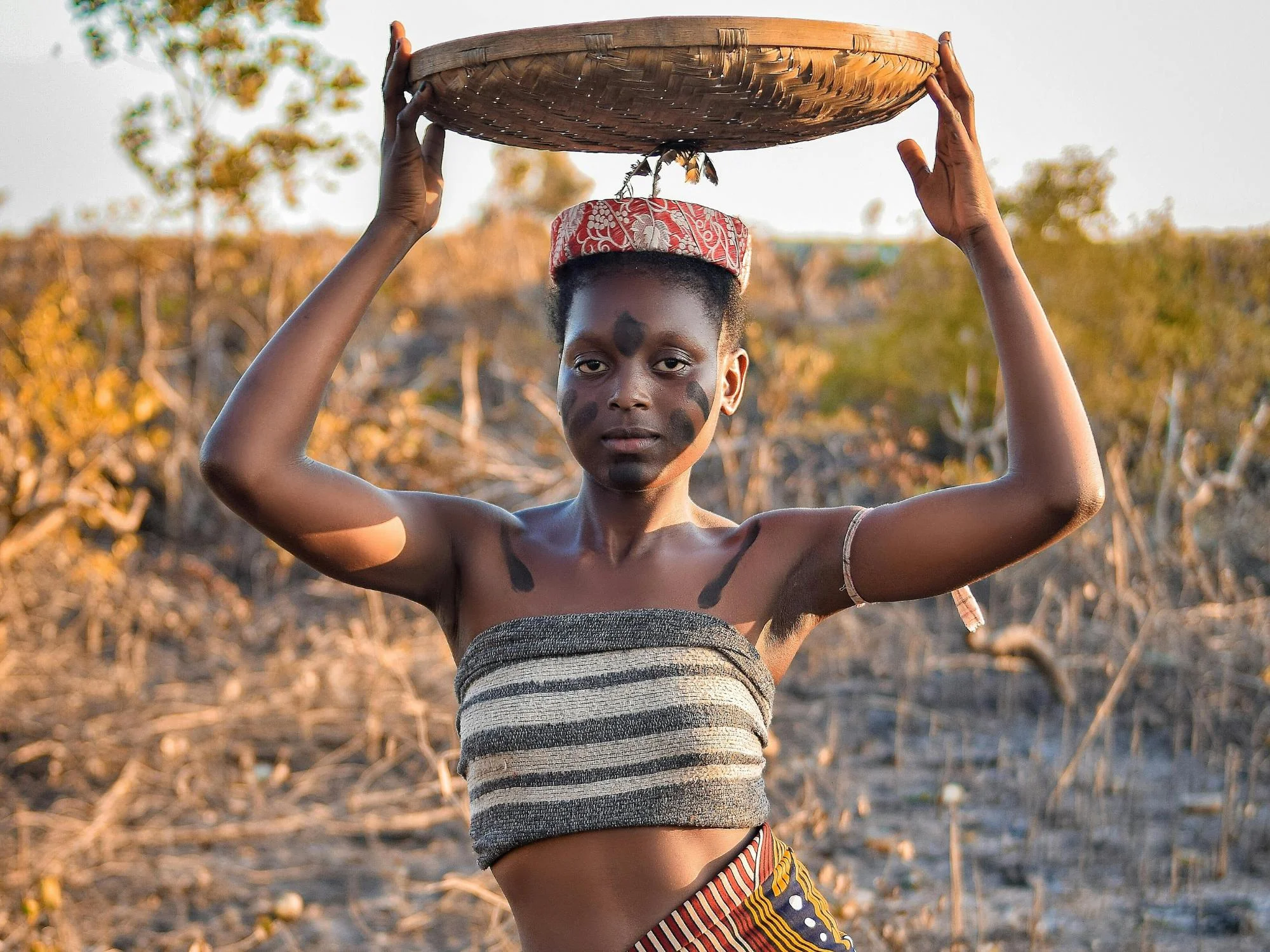Young Mozambican woman in traditional attire and face markings balancing a woven basket above her head