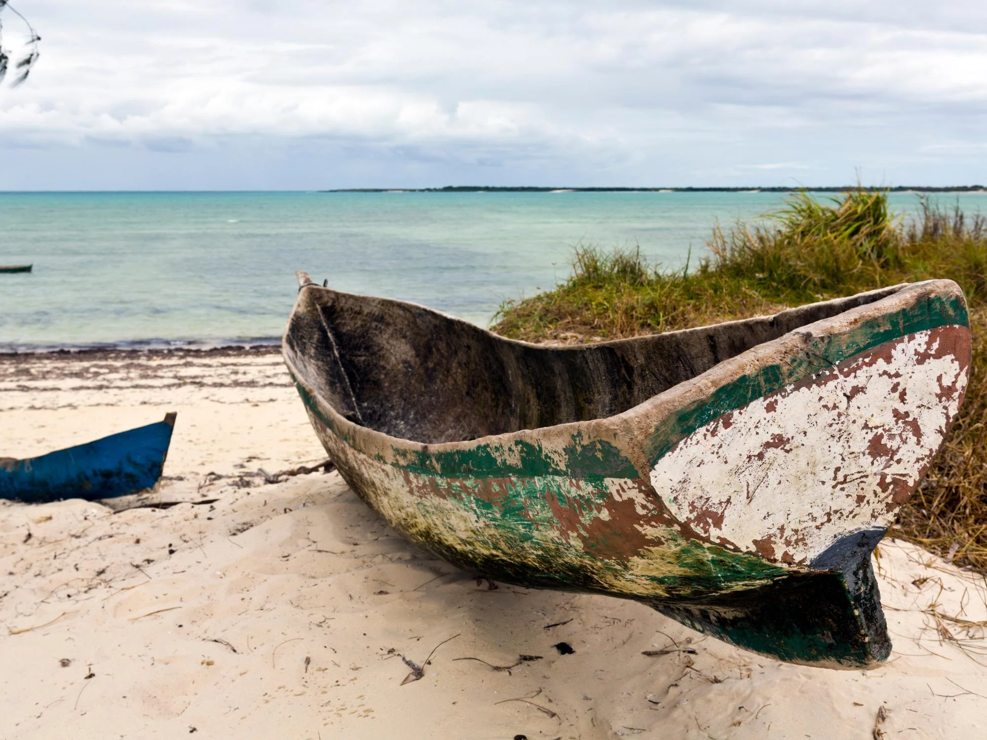 Close-up of a weathered dugout canoe with peeling paint resting on white sand beside turquoise water, Mozambique