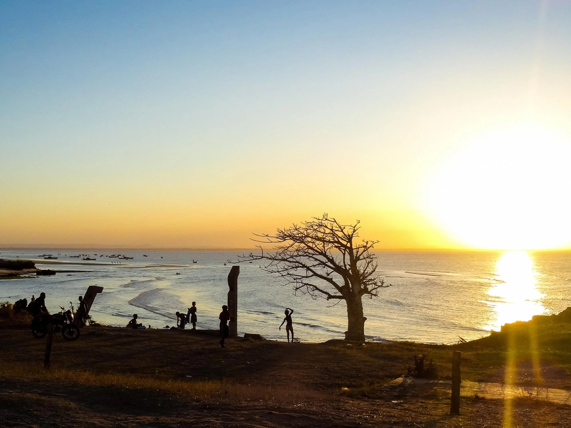 Silhouettes of local people and a bare tree against a vivid orange sunset over a Mozambican coastal bay