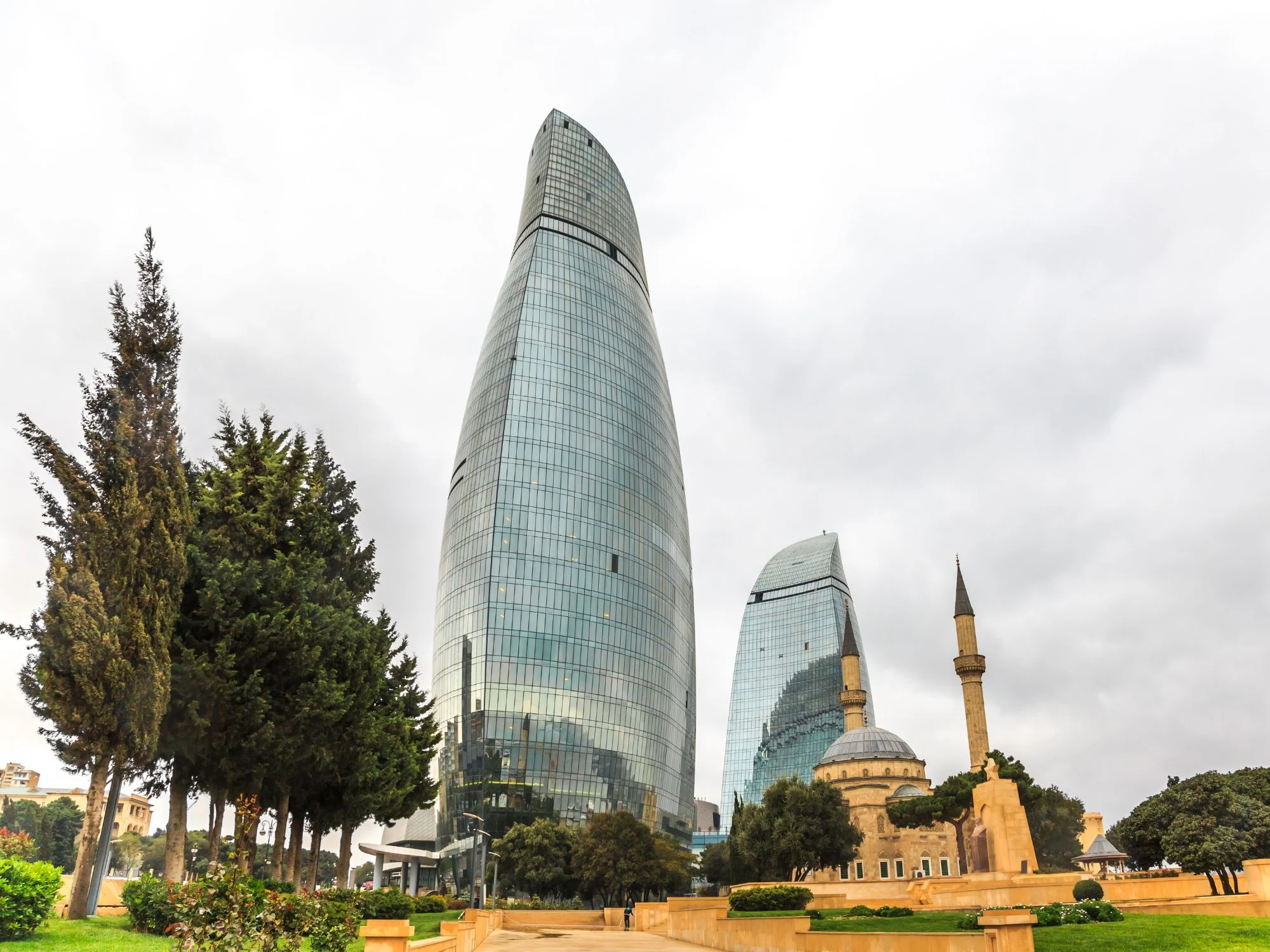 Flame Towers of Baku from Highland Park, with cypress trees and a small mosque minaret in the foreground
