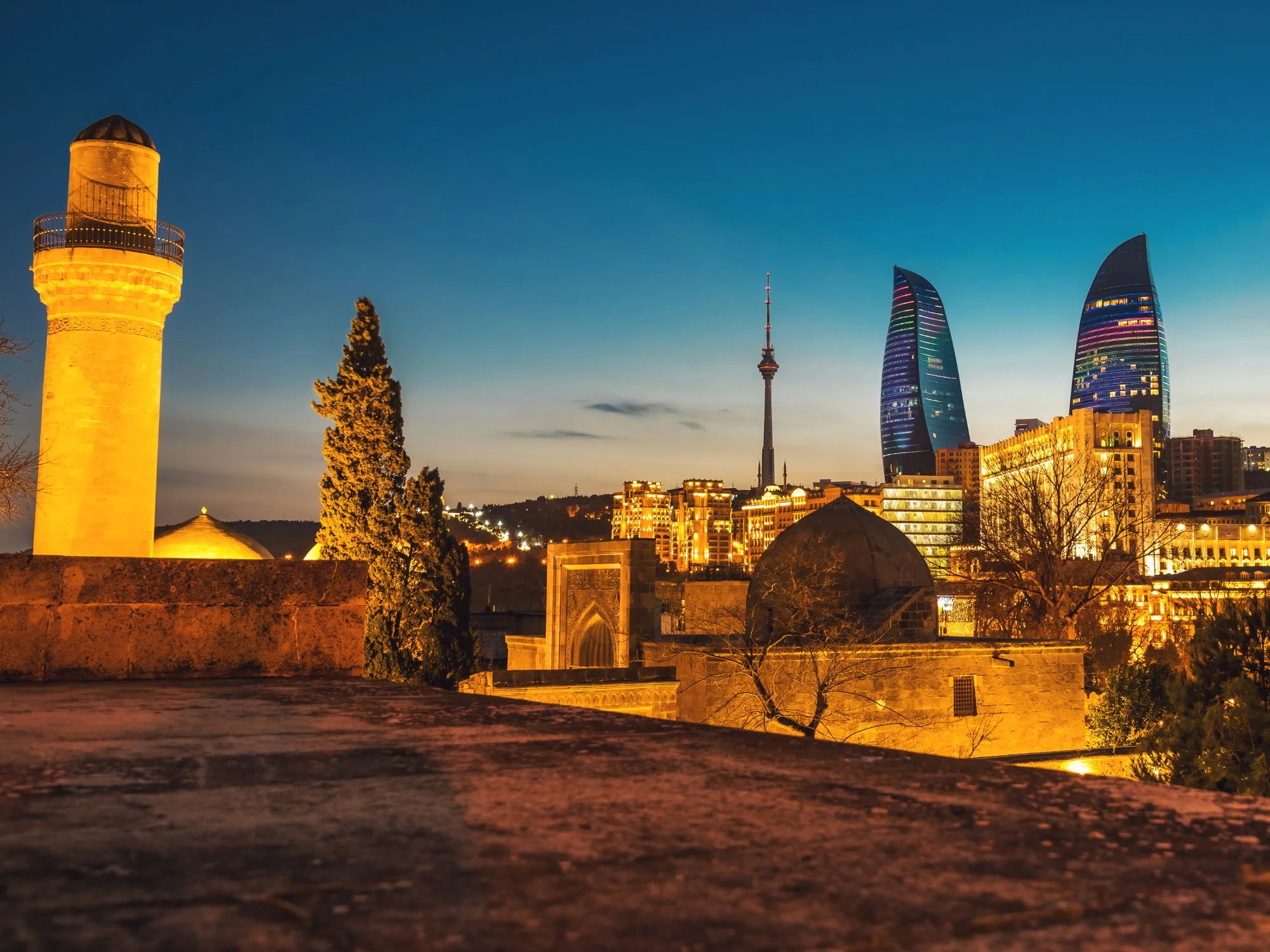 Twilight view of Baku — illuminated Old City watchtower in the foreground, Flame Towers and modern skyline beyond