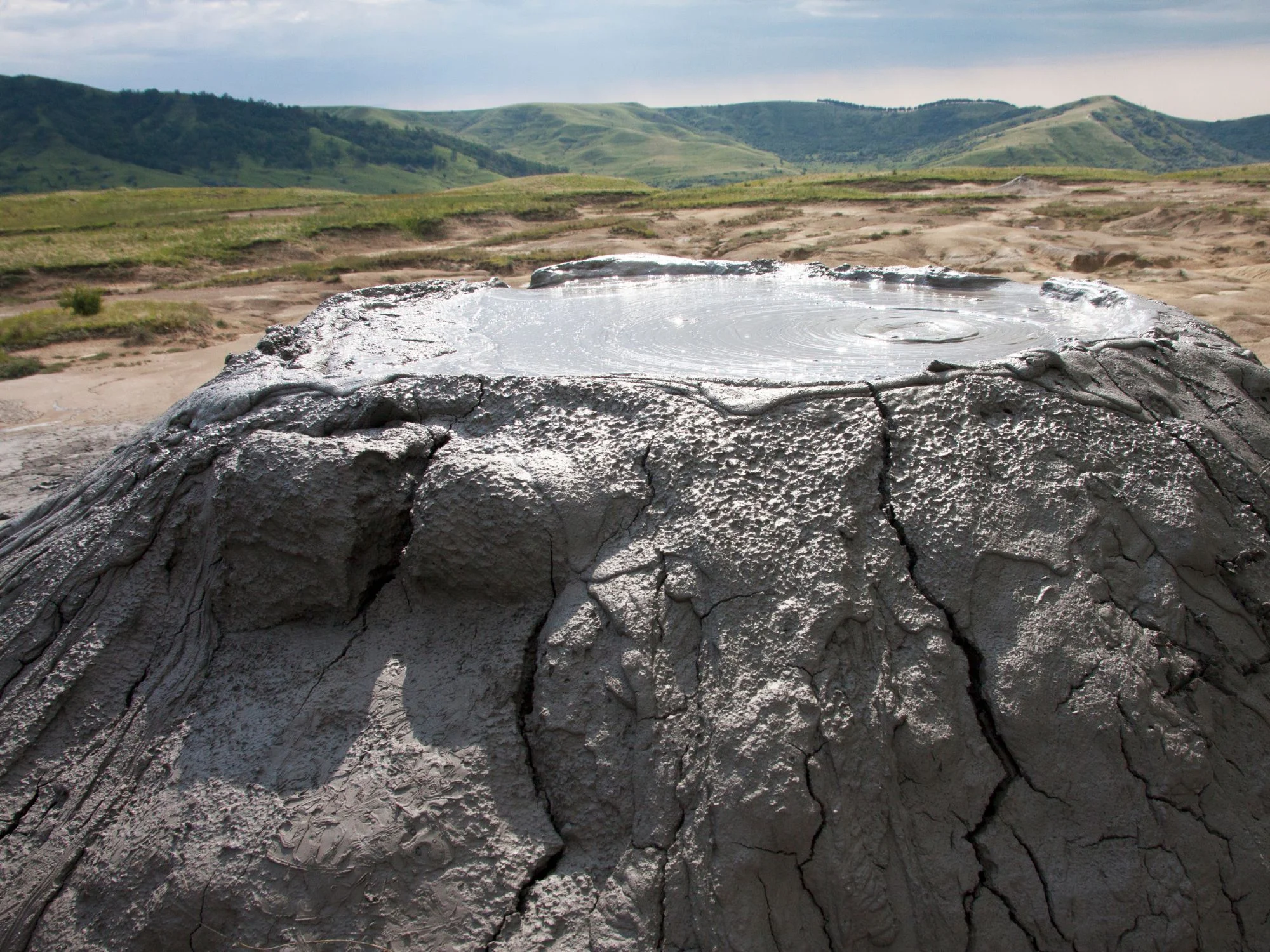 Bubbling mud volcano crater in Gobustan, Azerbaijan, with cracked grey terrain and rolling green hills behind