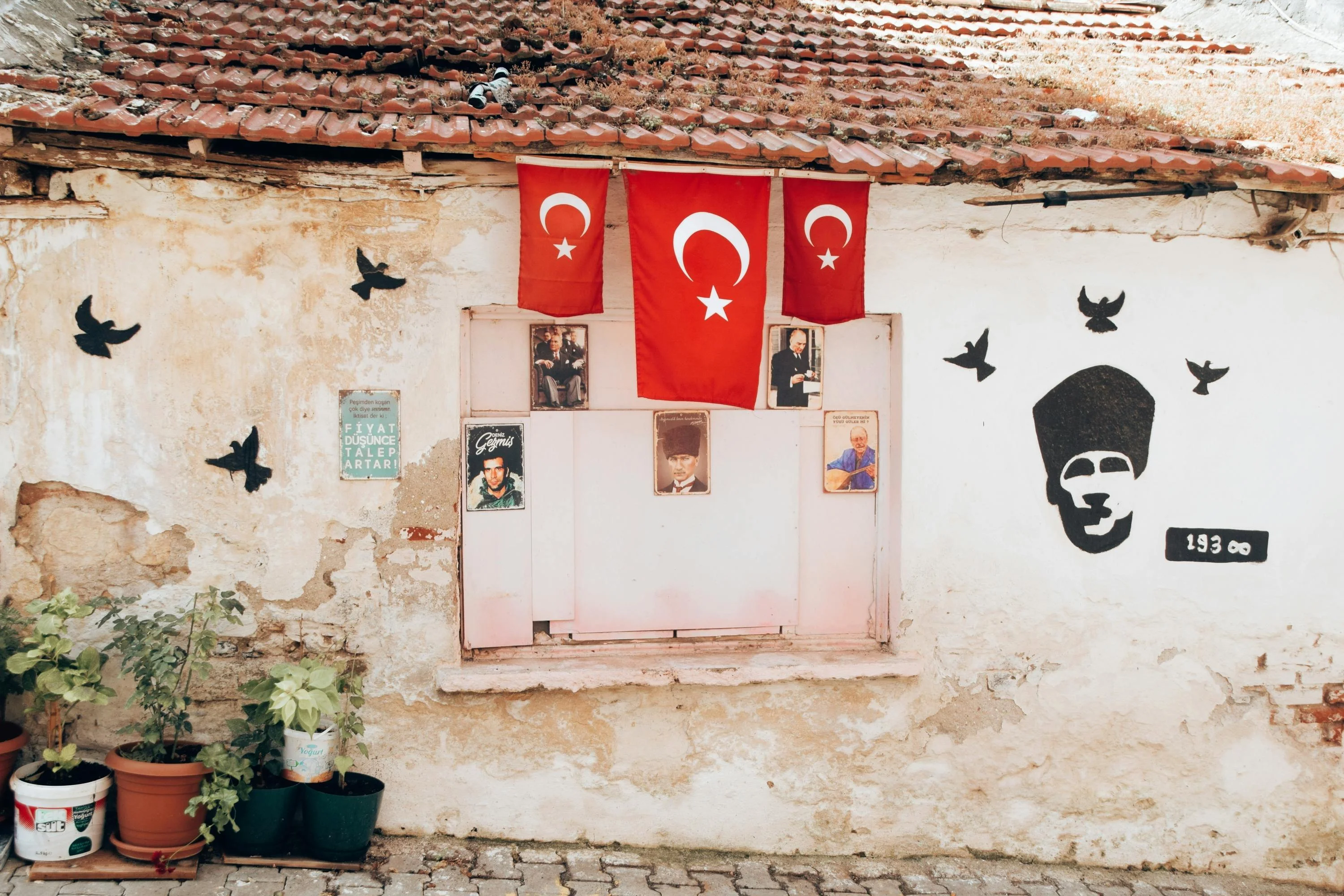 Weathered village wall with three Turkish flags, framed photographs and a stencilled portrait of Atatürk
