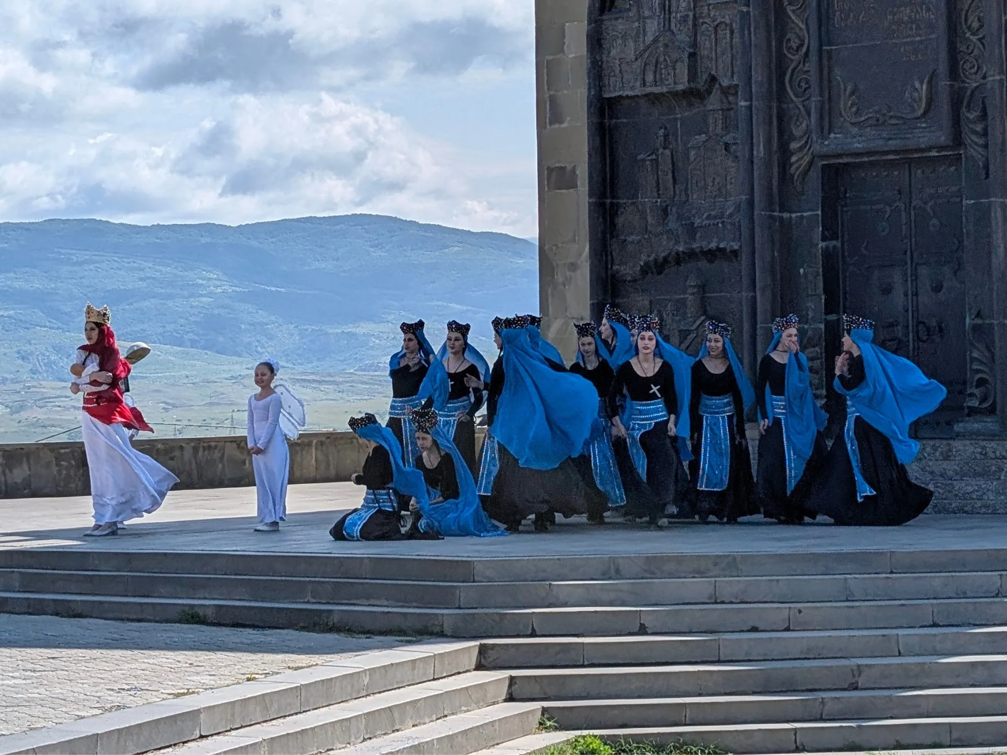 Folk dancers in long blue dresses and white veils performing in front of a stone church facade