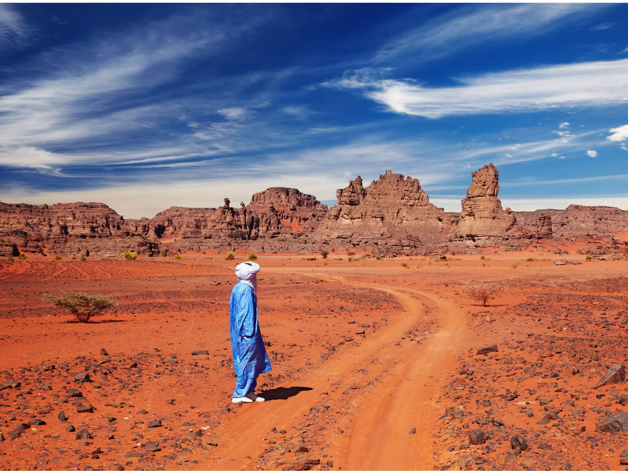 Tuareg man in traditional indigo robe on a red sand track among sandstone formations of the Sahara