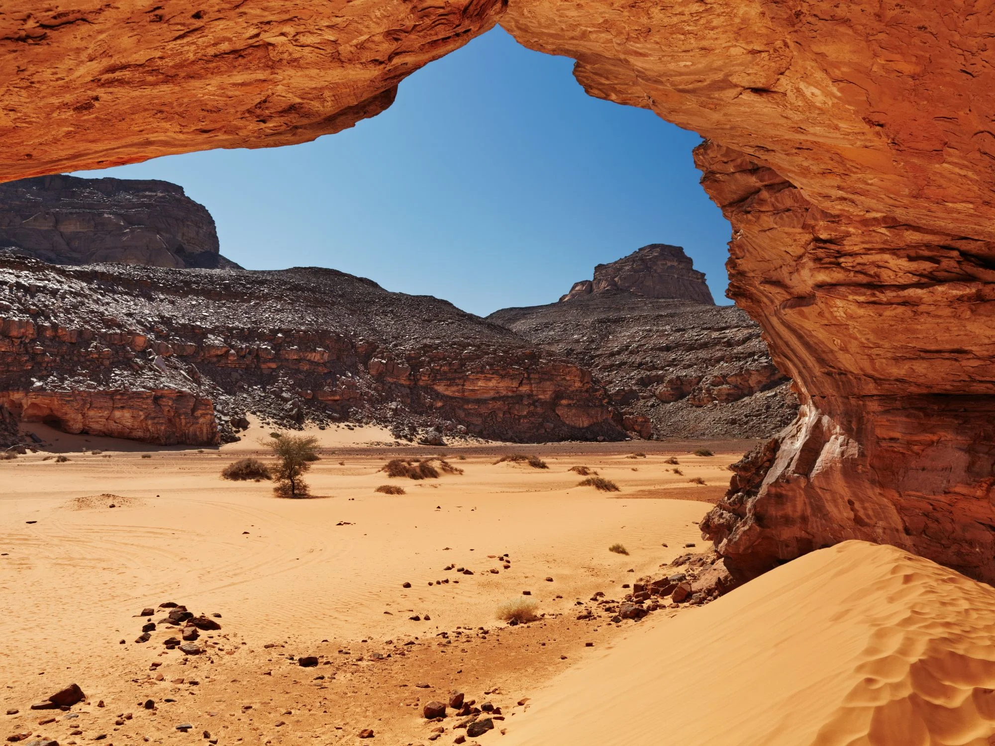 Natural sandstone rock arch framing a rugged Sahara landscape with dunes and dark mountains beyond