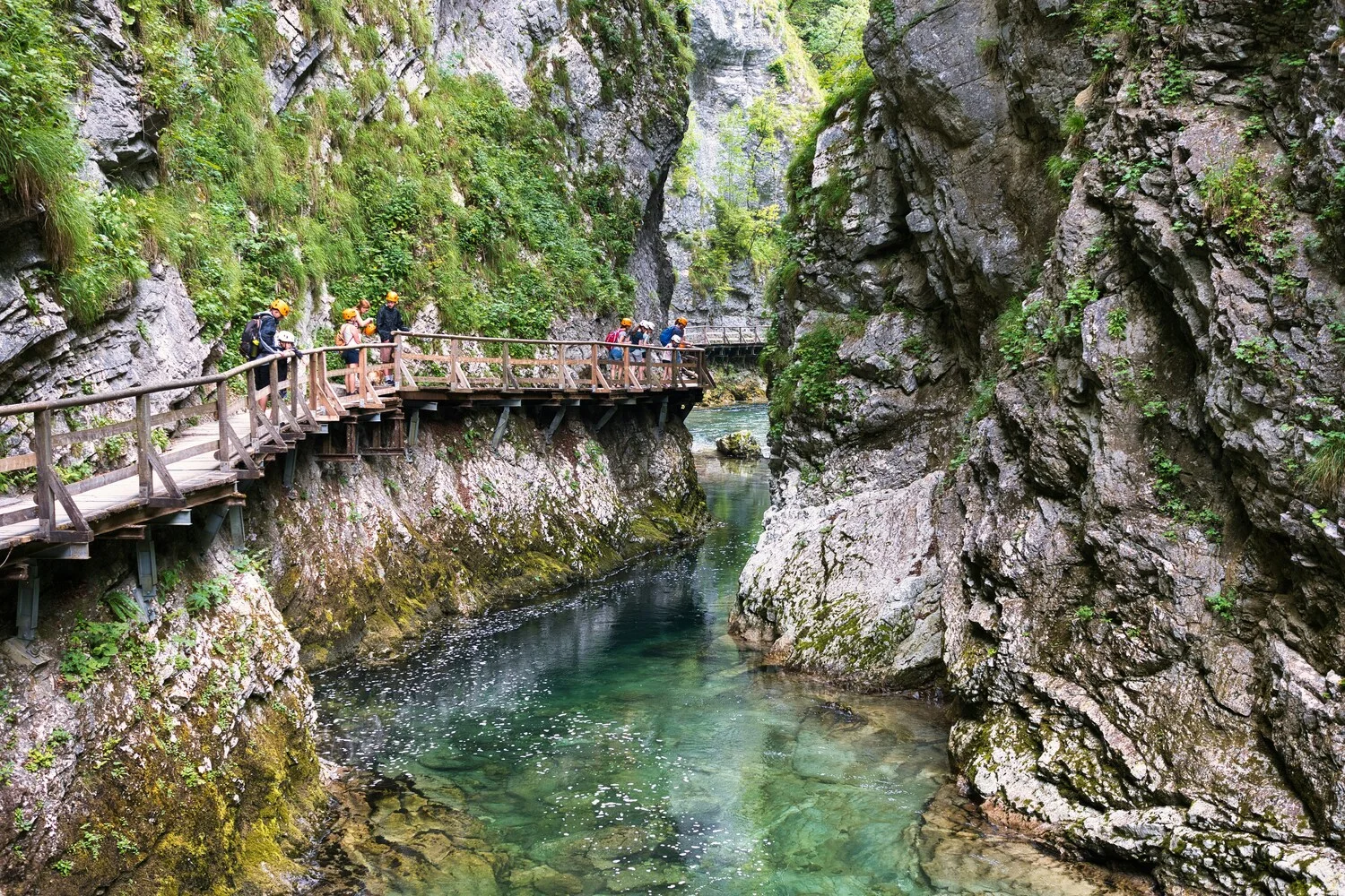Visitors in helmets crossing the wooden boardwalk above a clear turquoise pool in Vintgar Gorge, Slovenia