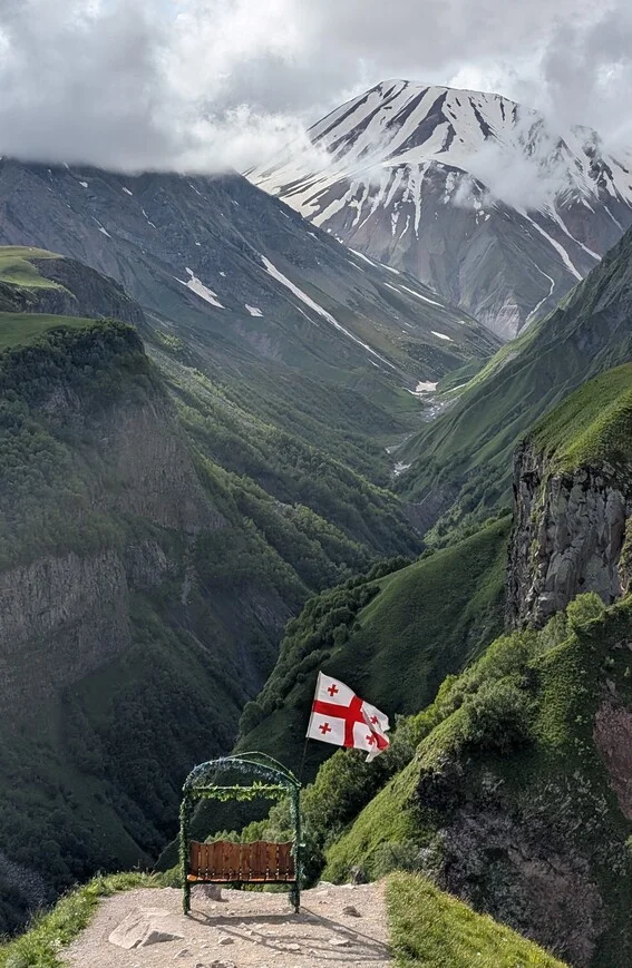 Bench draped with the Georgian flag overlooking a deep valley below snow-capped peaks near Stepantsminda