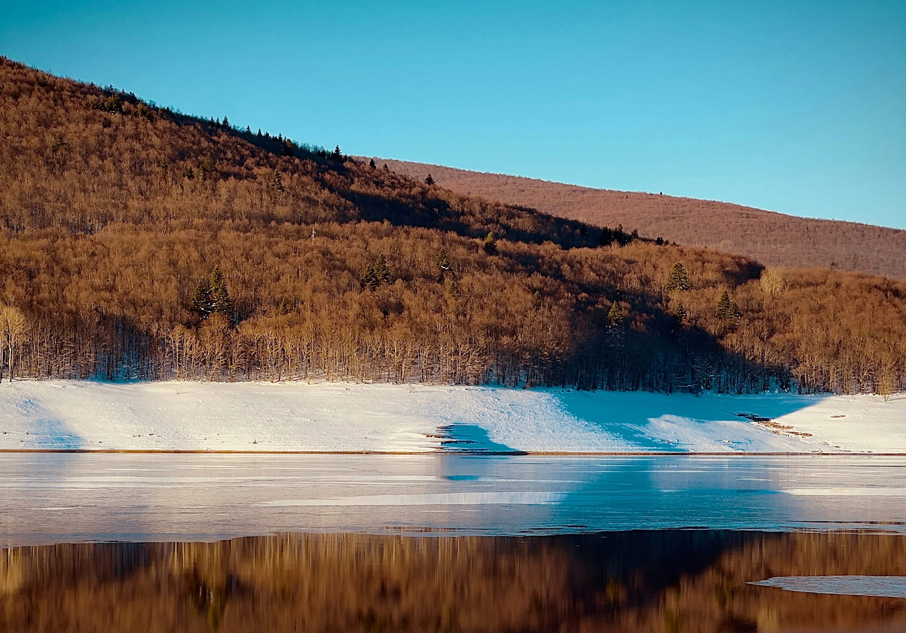 Frozen Mavrovo Lake reflecting a bare hillside forest at sunset in winter, North Macedonia