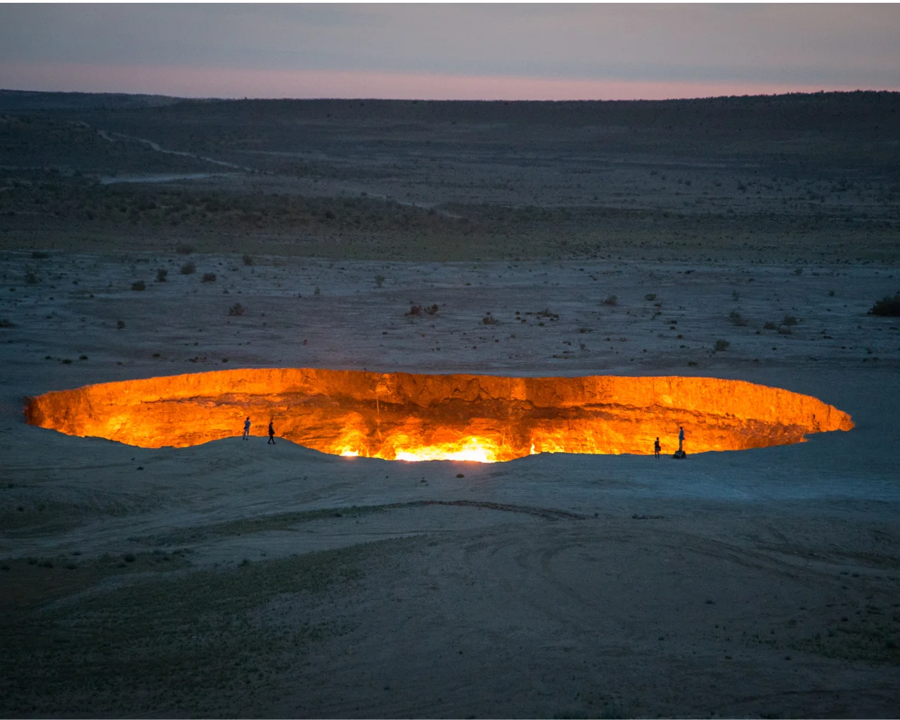 Glowing fiery crater of the Door to Hell at twilight with two tiny figures at the rim, Turkmenistan