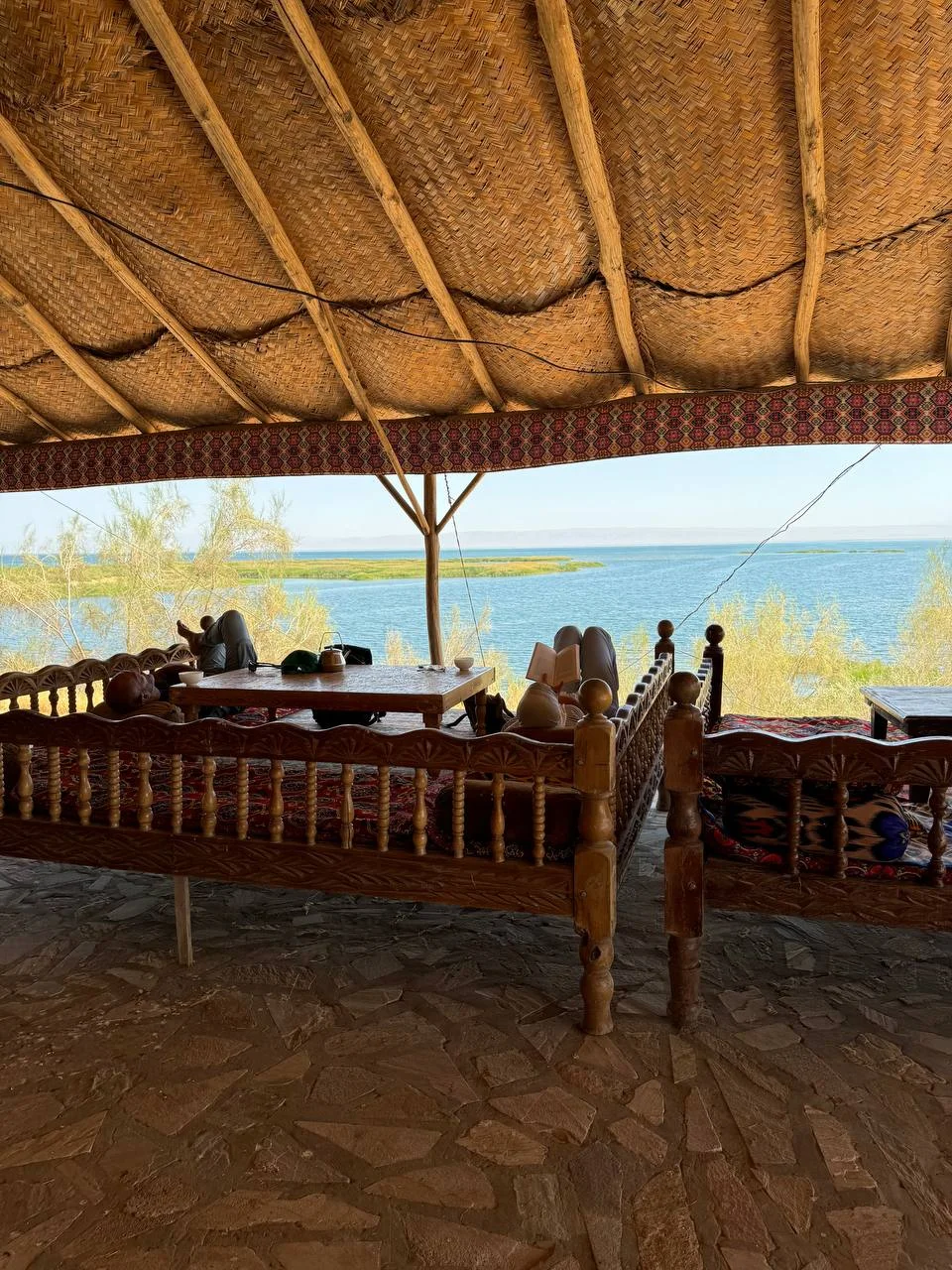 Carved wooden tapchans under a thatched-roof shelter overlooking the turquoise waters of Aydarkul Lake, Uzbekistan