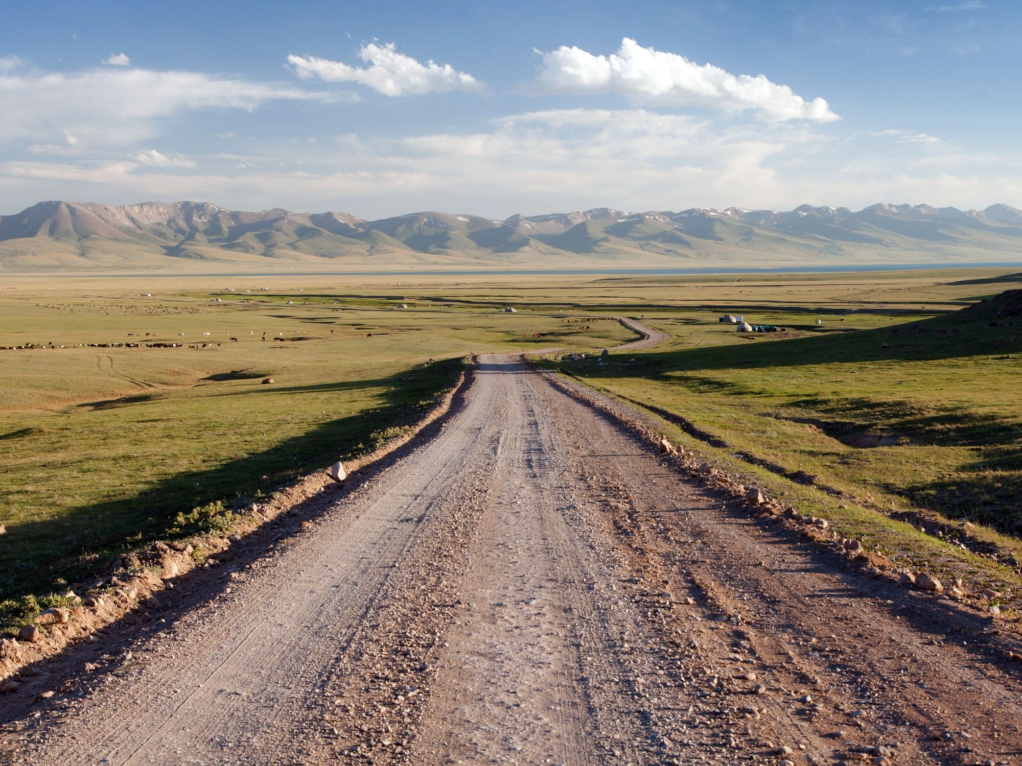 Dirt road through a wide green steppe with snow-streaked Tian Shan mountains and scattered yurts, Kyrgyzstan