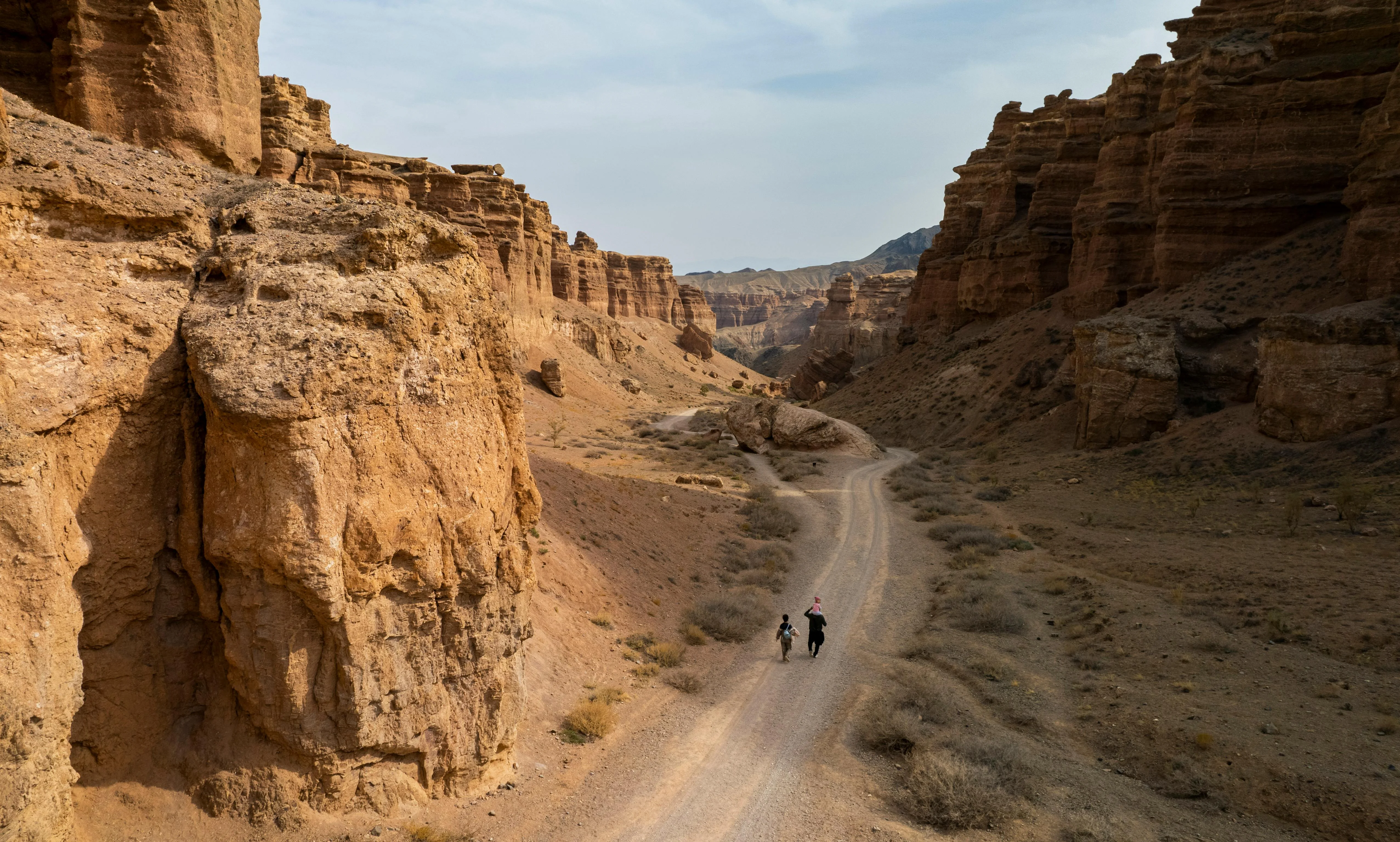 Two hikers walking the canyon-floor path between striated red sandstone cliffs in Charyn Canyon, Kazakhstan