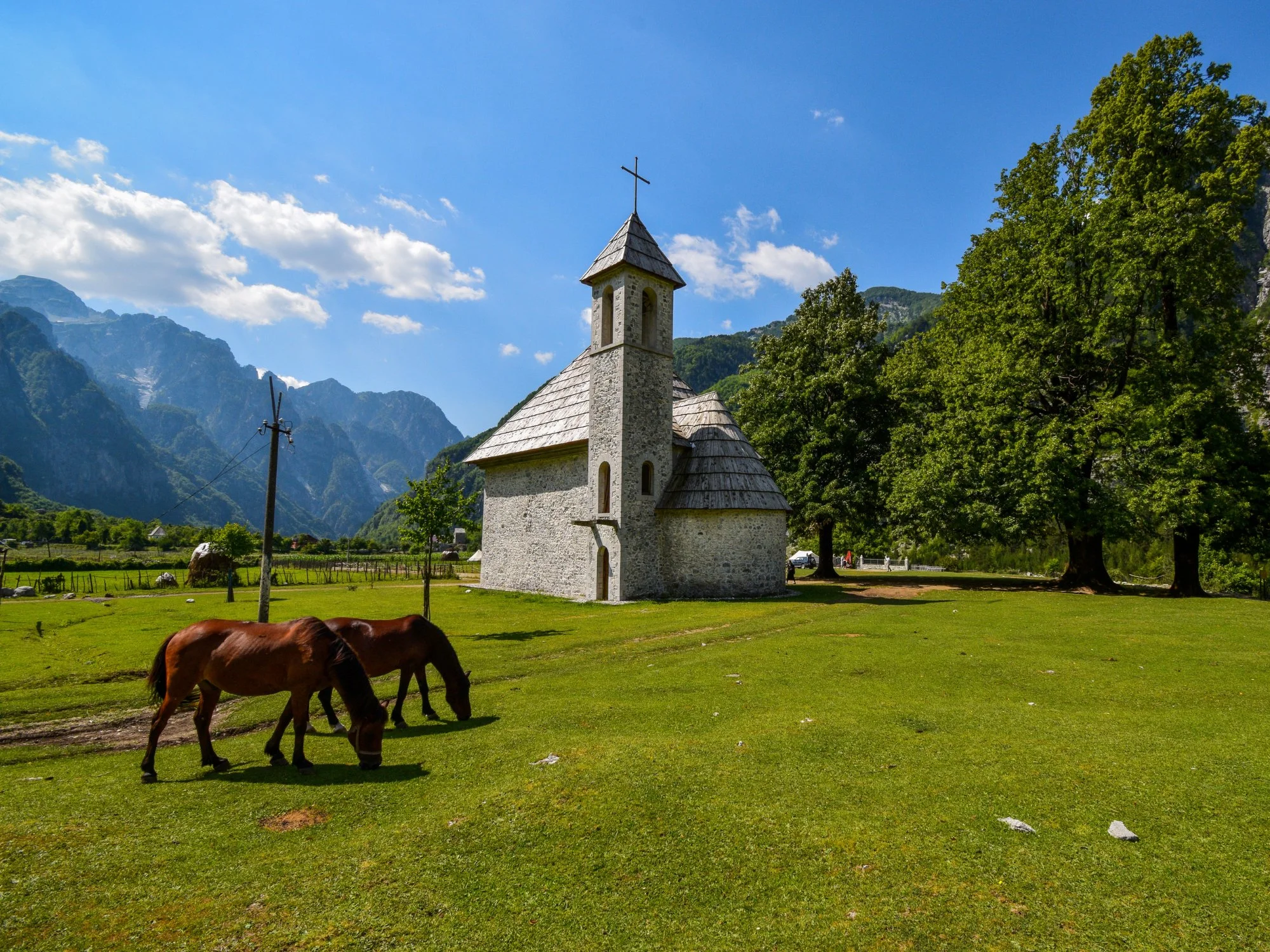 Stone Catholic church and grazing horses in Theth village beneath the snowcapped Albanian Alps