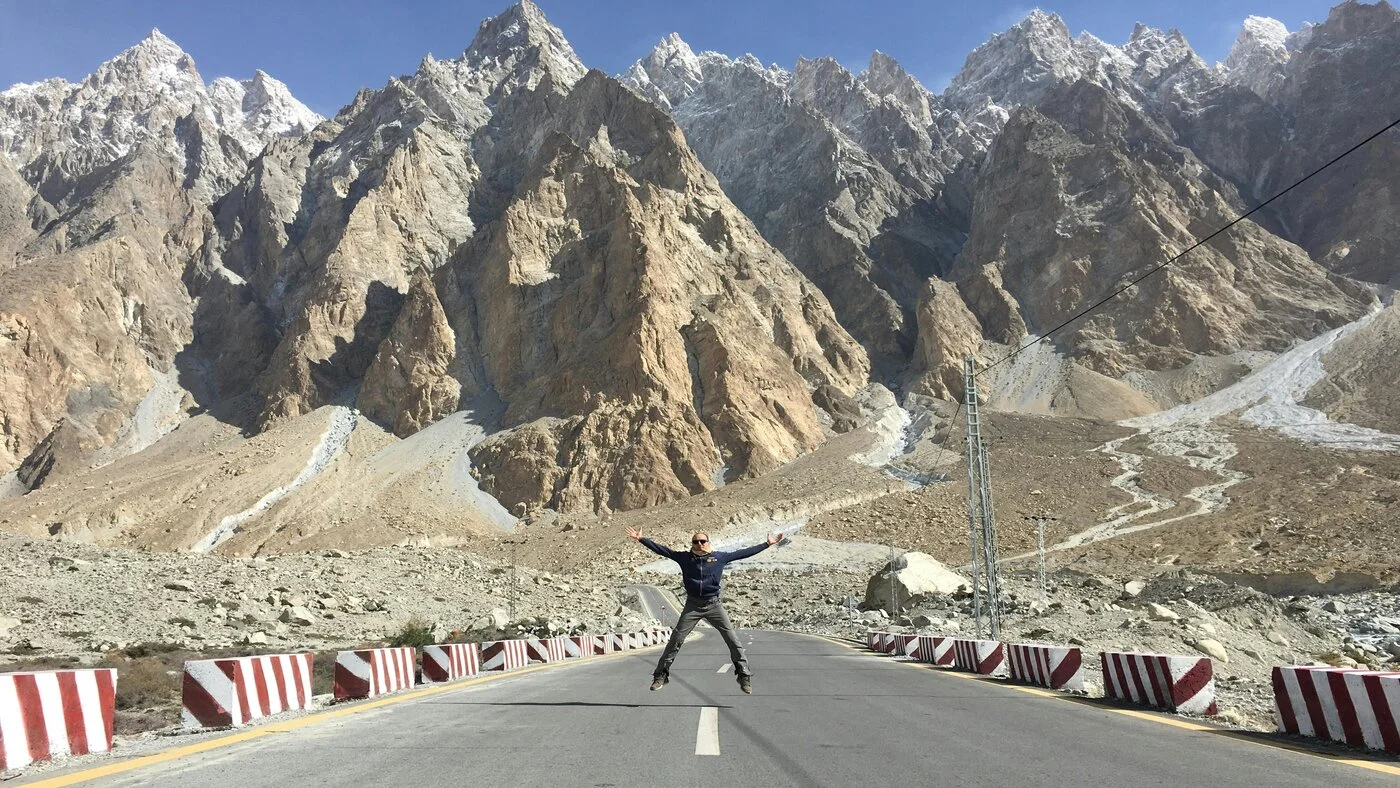 Traveler with arms raised on the Karakoram Highway beneath the jagged Passu Cones, Hunza Valley, Pakistan