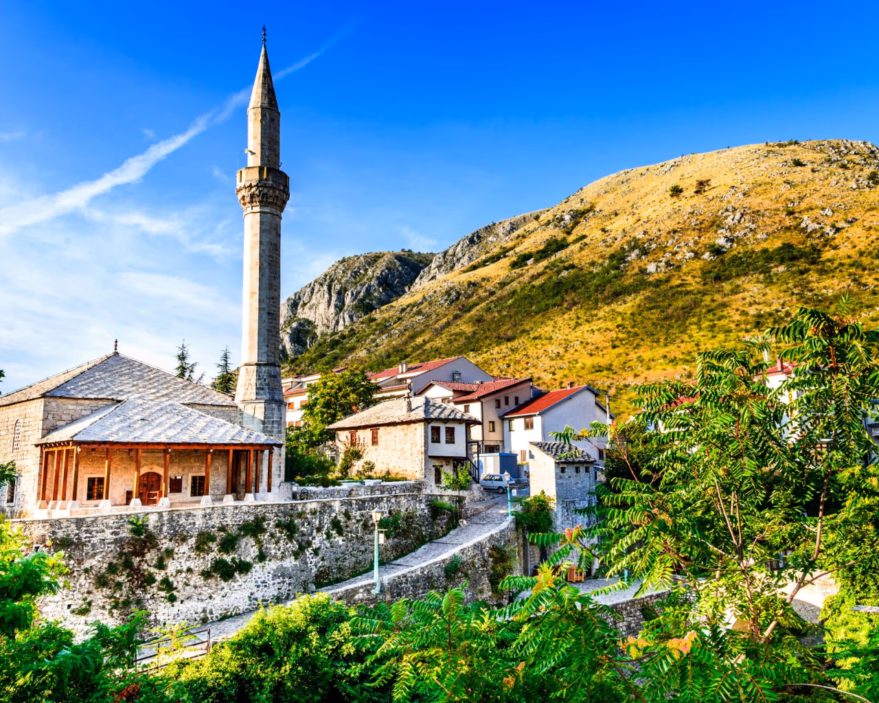 Rustic stone mosque with slender minaret nestled in a hillside village surrounded by greenery in the Balkans.
