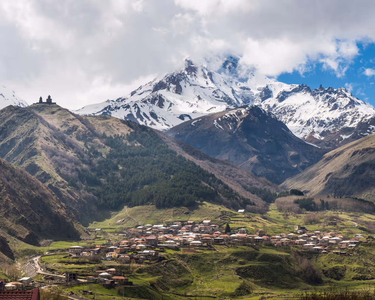 Snow-capped Mount Kazbegi with the Gergeti Trinity Church perched on a hill, overlooking a small village.