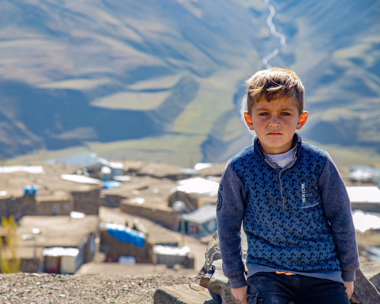 A young boy in Khinaliq, Azerbaijan, sitting on a rock with a stunning mountain village in the background.