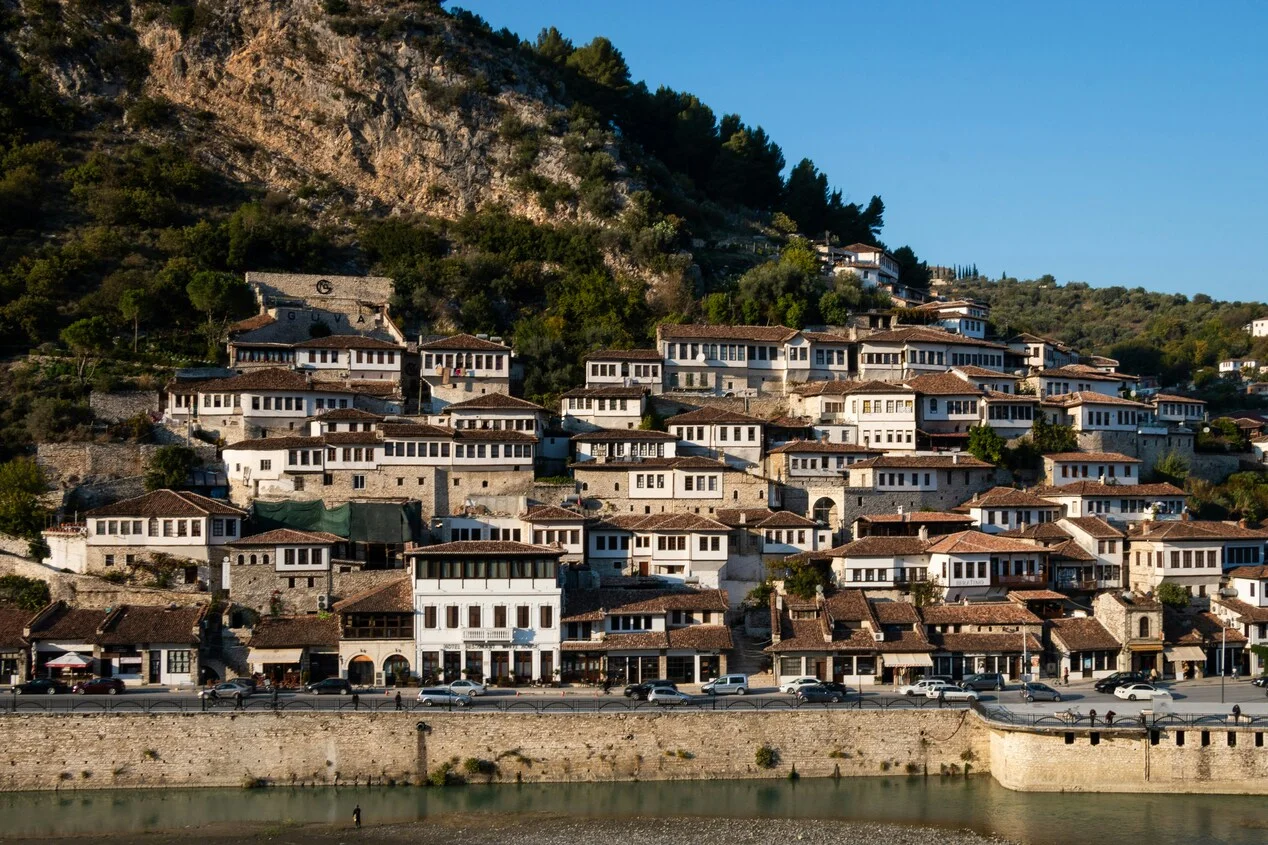 Panoramic view of Berat, Albania, with Ottoman-era white houses and red-tiled roofs climbing the hillside above the Osum River beneath a rocky mountain backdrop.