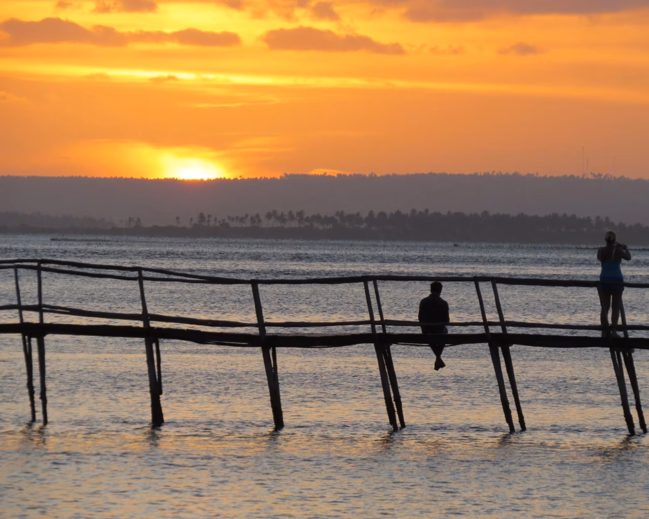 Two silhouetted figures on a wooden pier watching a brilliant orange sunset over a Mozambican bay