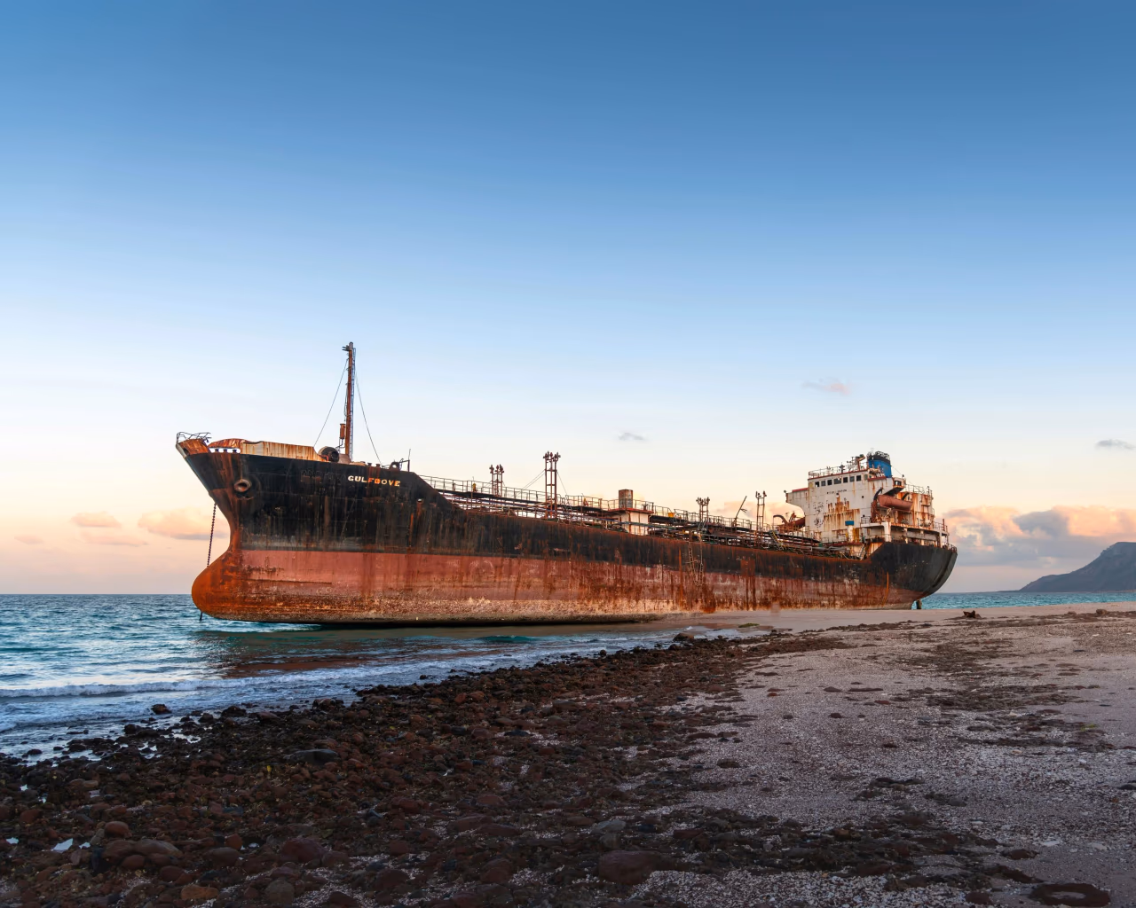 Rusting cargo shipwreck stranded on Socotra shore with turquoise sea and mountains in background.