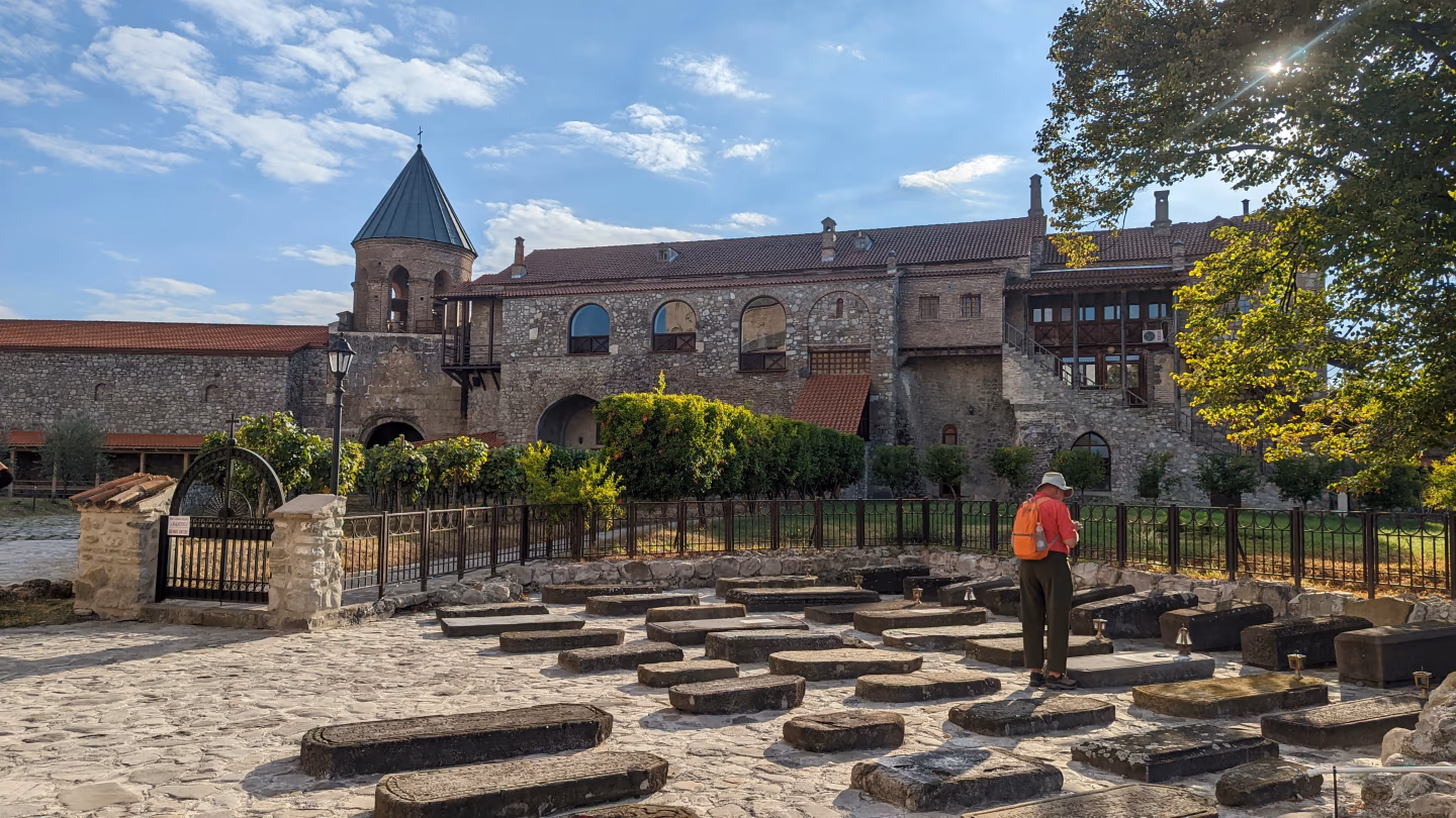 Georgian monastery cemetery – traveler walking among old stone graves at a historic monastery complex with bell tower and red tiled roofs.