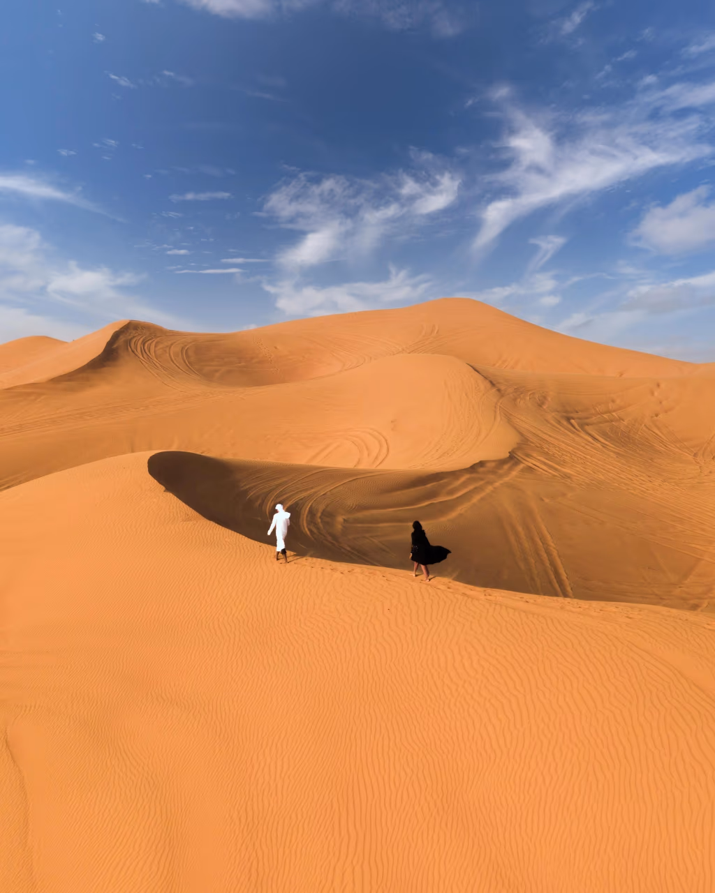 Two people walking across tall desert dunes under blue sky