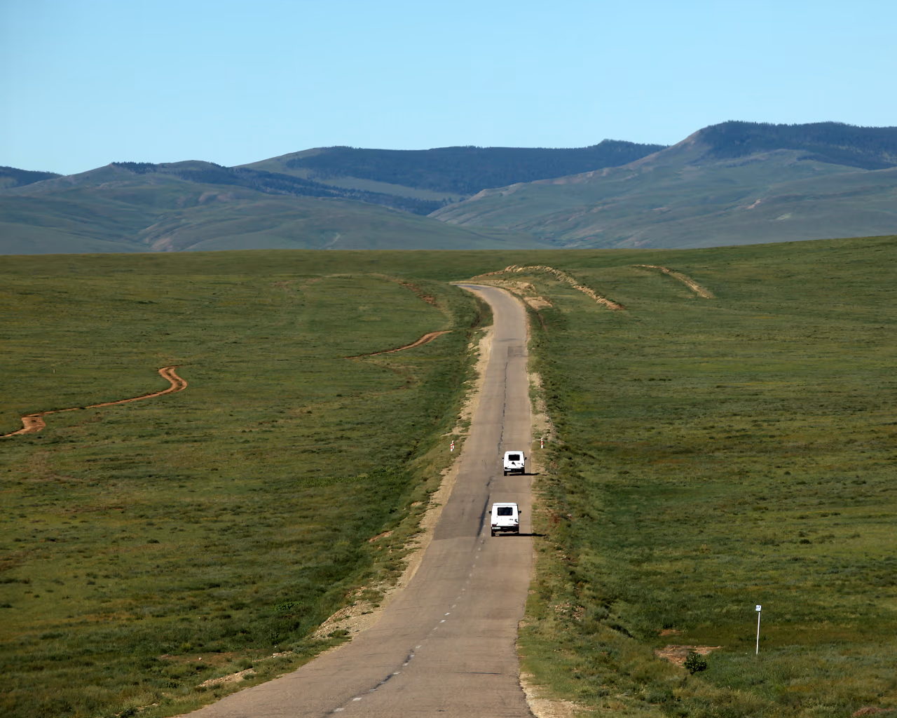 Long paved road crossing Mongolia grasslands with green hills and white vans driving ahead.