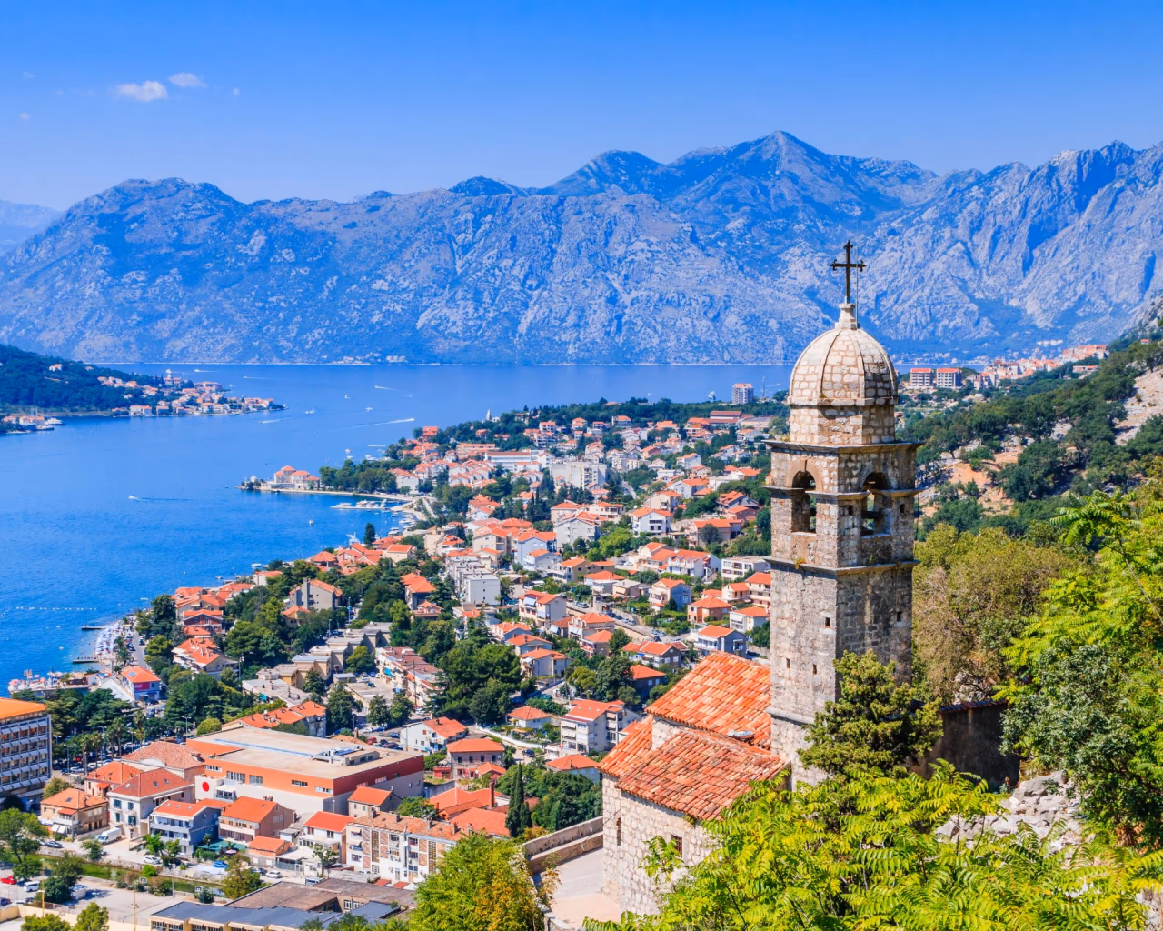 Scenic view of Kotor Bay and Old Town rooftops with a stone church tower, surrounded by mountains, Montenegro.