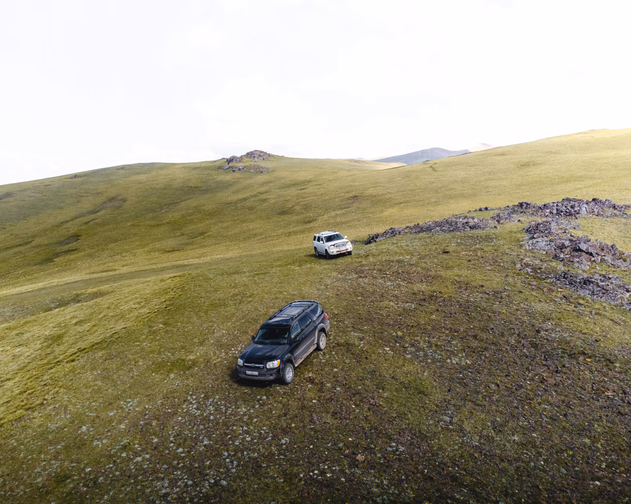 Two off-road vehicles parked on grassy Kyrgyz plateau surrounded by rolling hills.