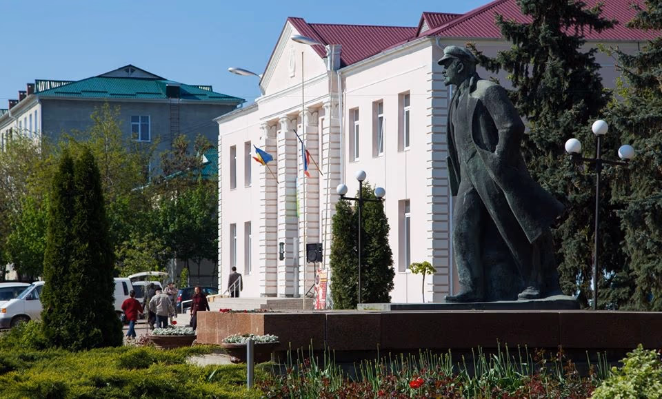 Lenin statue in Comrat, Moldova, near administrative building with flags and red roof.