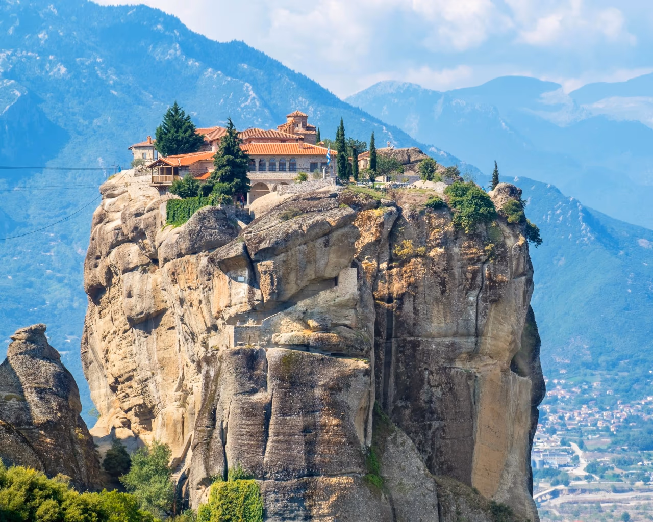 Monastery perched on massive rock cliff in Meteora Greece with blue mountains in background.