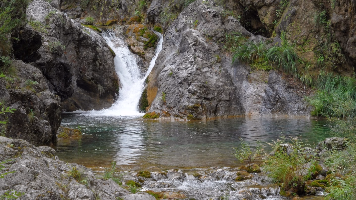 Small waterfall cascading into a turquoise pool, surrounded by mossy rocks in Enippeus Gorge, Mount Olympus, Greece.